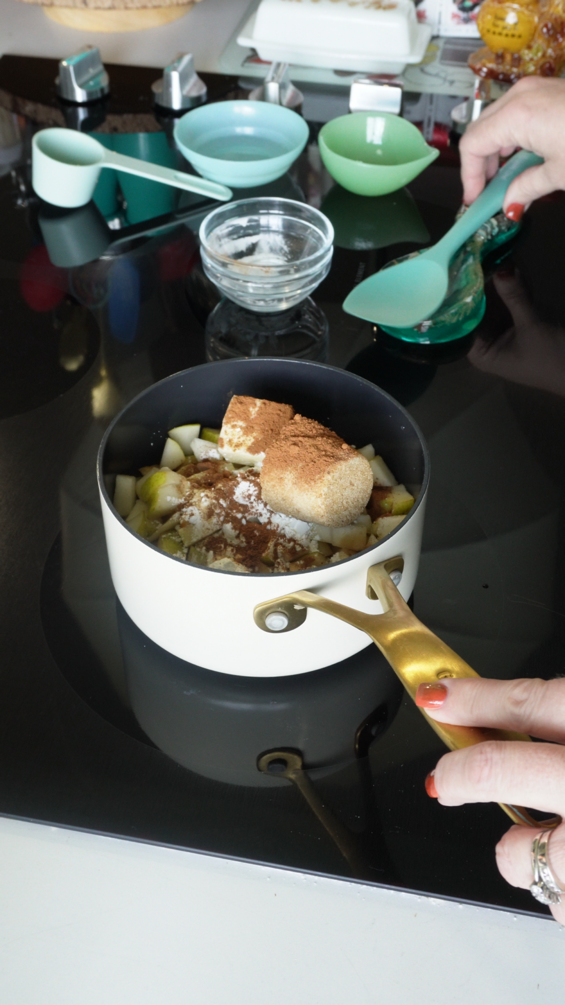 A person adds brown sugar and cinnamon to diced apples in a white pot on a black stove. Measuring cups and bowls are nearby.