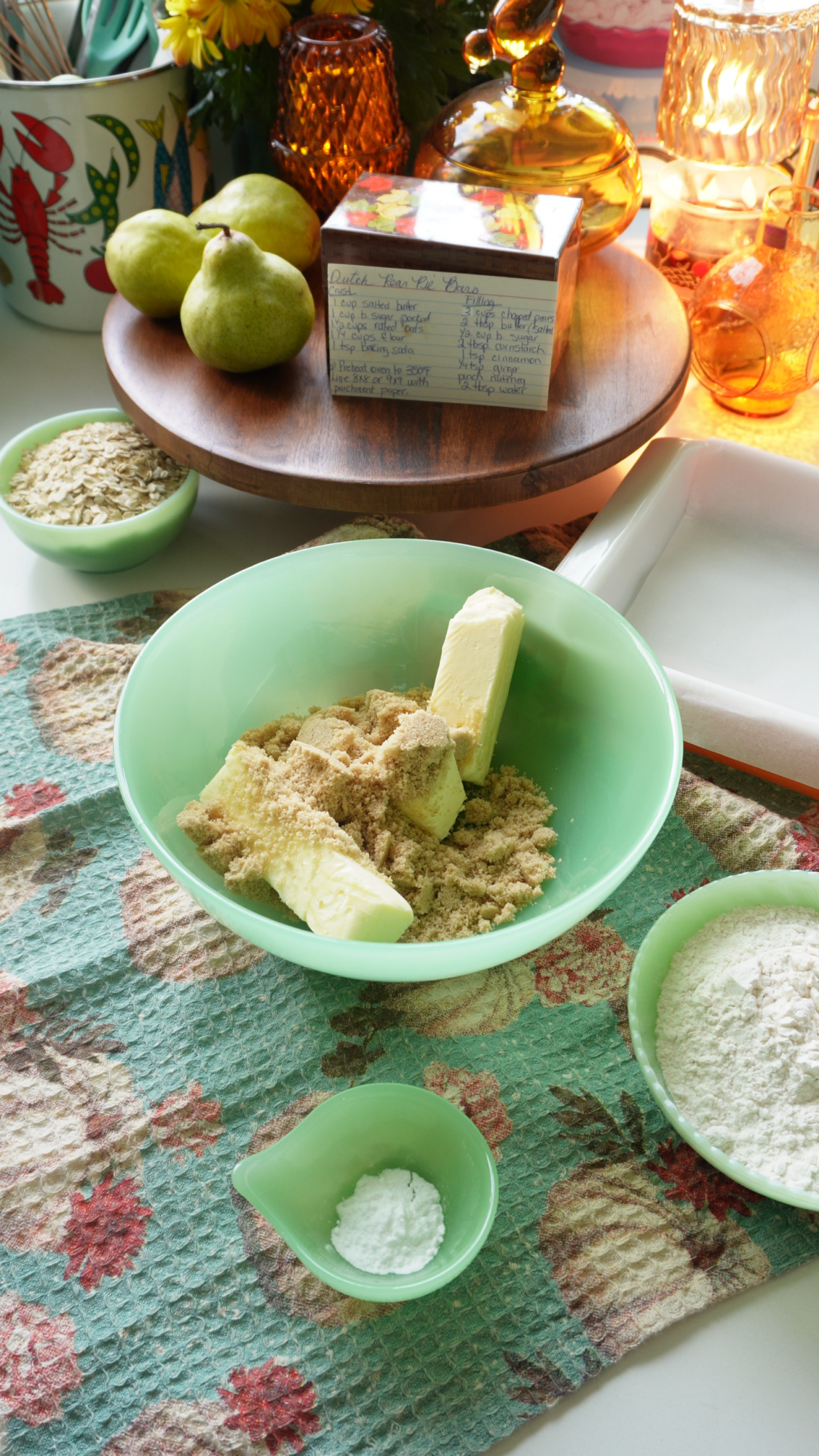 A green bowl with butter and brown sugar sits on a floral towel, surrounded by flour, baking powder, green apples, oats, and vintage glassware. A handwritten recipe card is on a wooden tray nearby.