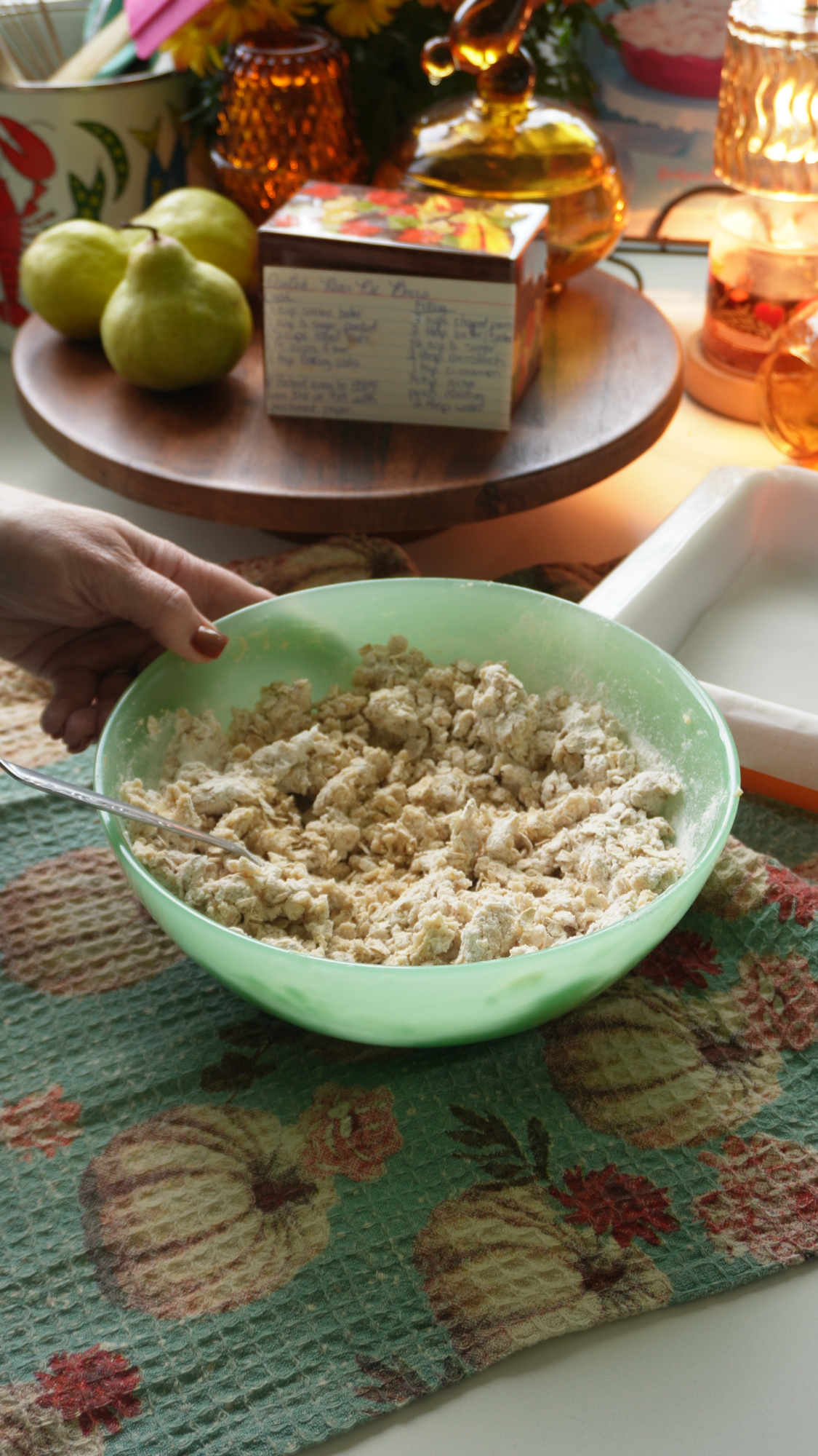 A hand holds a spoon in a green bowl filled with dough. Nearby are pears, a recipe card, and fall-themed decor on a pumpkin-patterned towel. Warm lighting creates a cozy kitchen atmosphere.