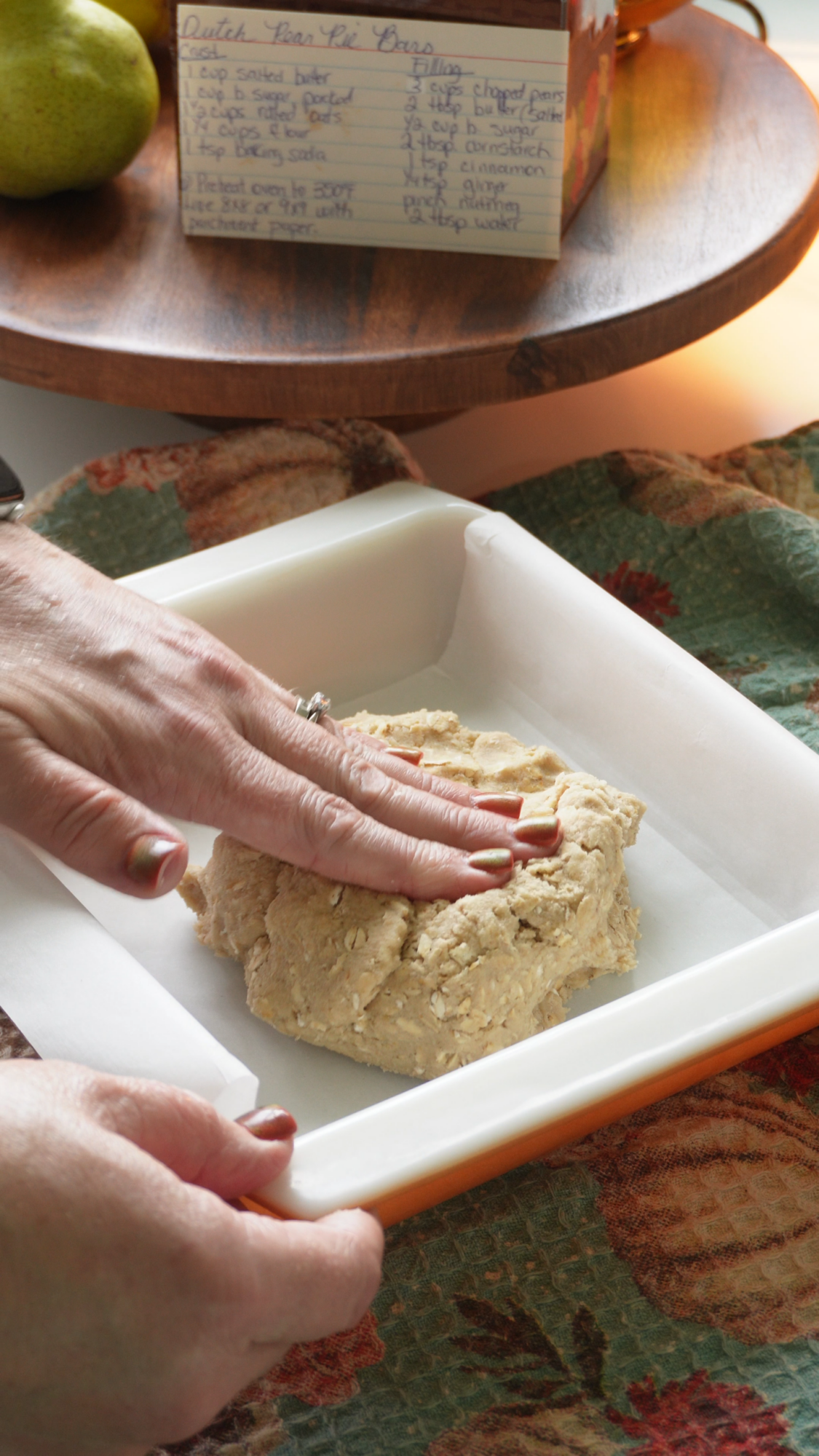 A person presses dough into a parchment-lined baking dish next to a handwritten recipe card and a decorative autumn cloth.