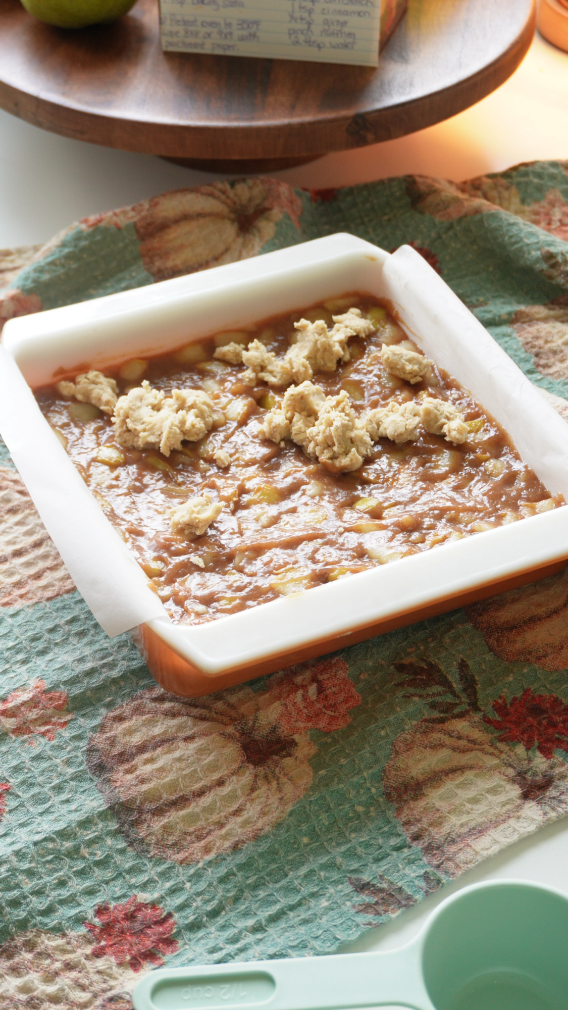 A baking dish with an unbaked dessert topped with clumps of dough sits on a pumpkin-patterned towel, next to a green measuring cup.
