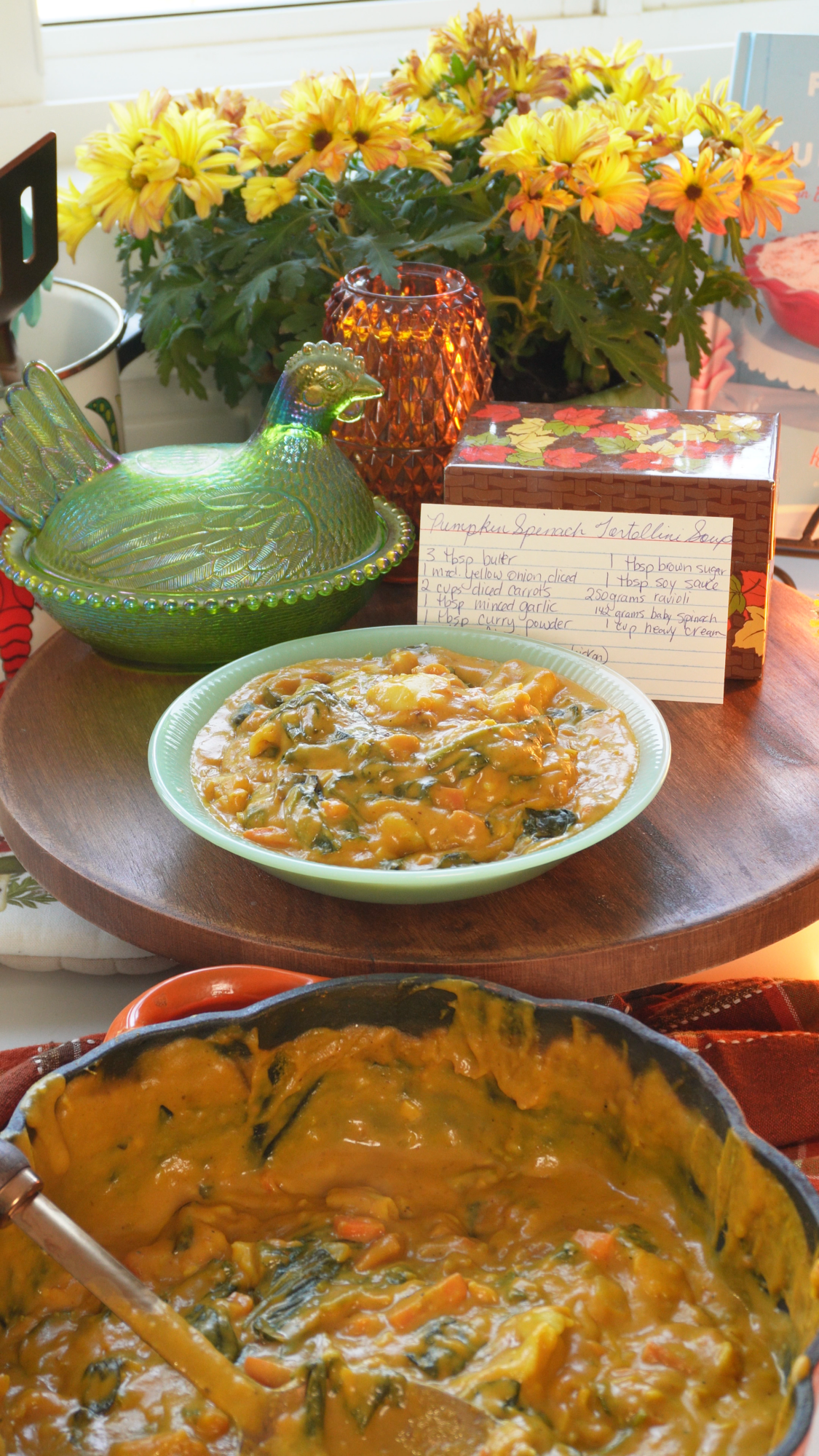 A bowl of thick pumpkin spinach tortellini soup sits on a table with flowers, a green ceramic hen, a recipe card, and a pot filled with more soup in the foreground.