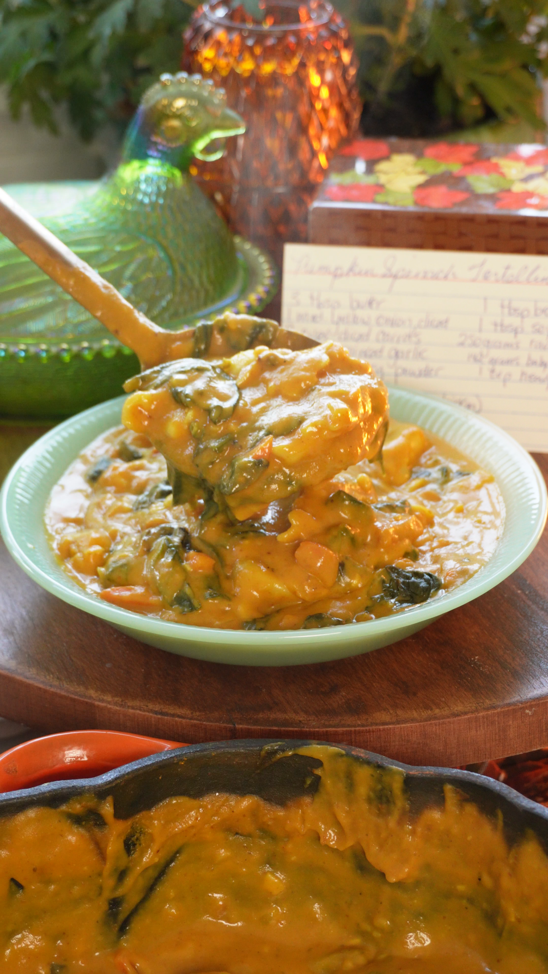 A spoon lifts creamy pumpkin and spinach tortellini from a green bowl, surrounded by autumn decor and a handwritten recipe card in the background.