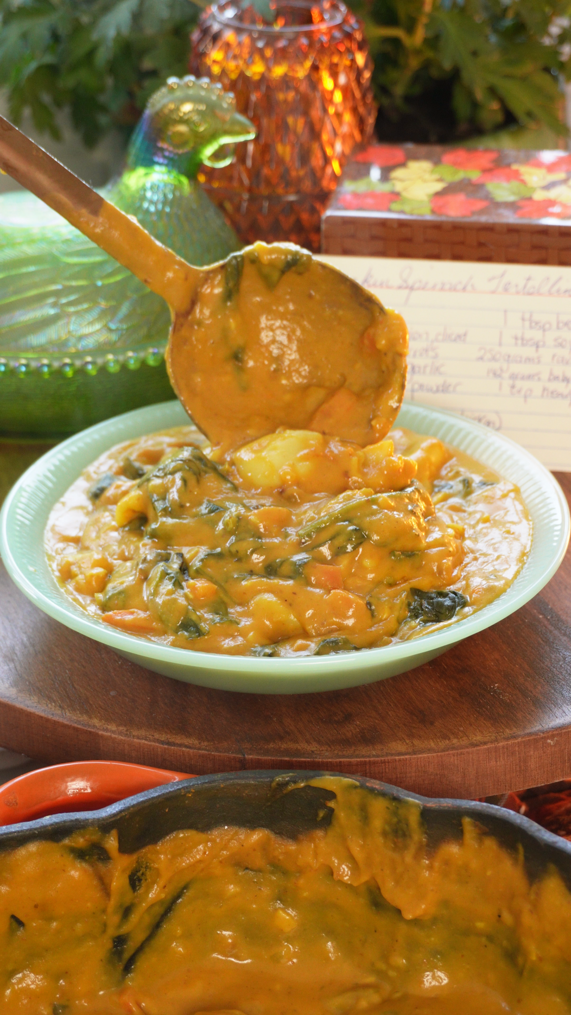 A ladle lifts creamy, orange-hued spinach tortellini from a light green bowl. A recipe card sits in the background, near a decorative glass hen and amber vase, while a cast-iron skillet of the same dish sits in the foreground.