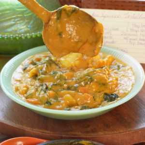 A ladle lifts creamy, orange-hued spinach tortellini from a light green bowl. A recipe card sits in the background, near a decorative glass hen and amber vase, while a cast-iron skillet of the same dish sits in the foreground.