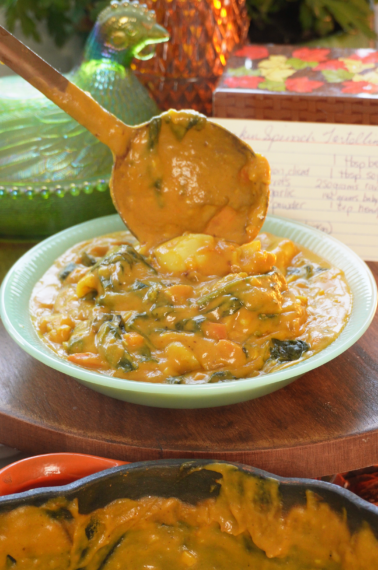 A ladle lifts creamy, orange-hued spinach tortellini from a light green bowl. A recipe card sits in the background, near a decorative glass hen and amber vase, while a cast-iron skillet of the same dish sits in the foreground.