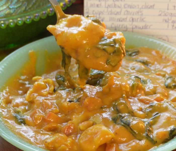A spoon lifts a serving of creamy, orange-yellow soup with spinach and vegetables from a bowl. In the background, a handwritten recipe card is partially visible.