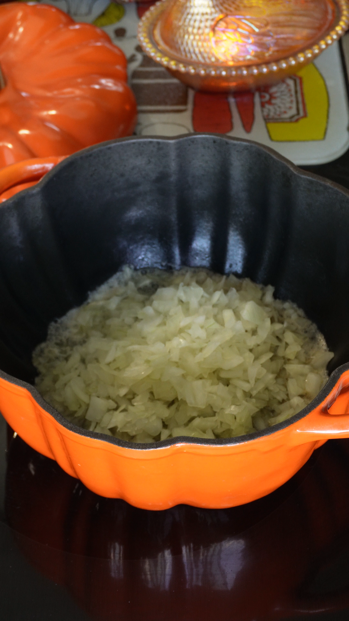 Chopped onions cooking in a vibrant orange, pumpkin-shaped pot on a stovetop. Other orange and glass kitchen items are visible in the background.