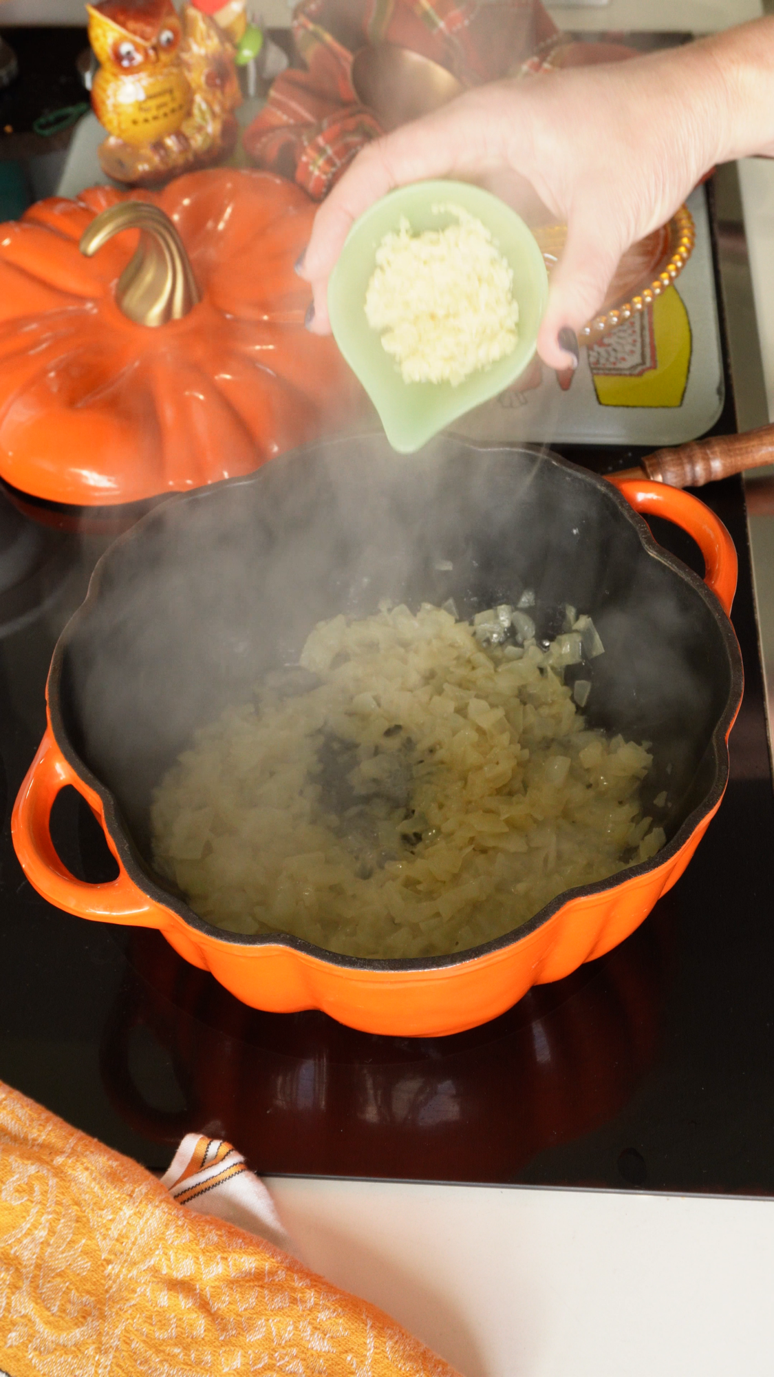A hand adds minced garlic from a green bowl into a steaming orange pan with sautéed onions on a stovetop.
