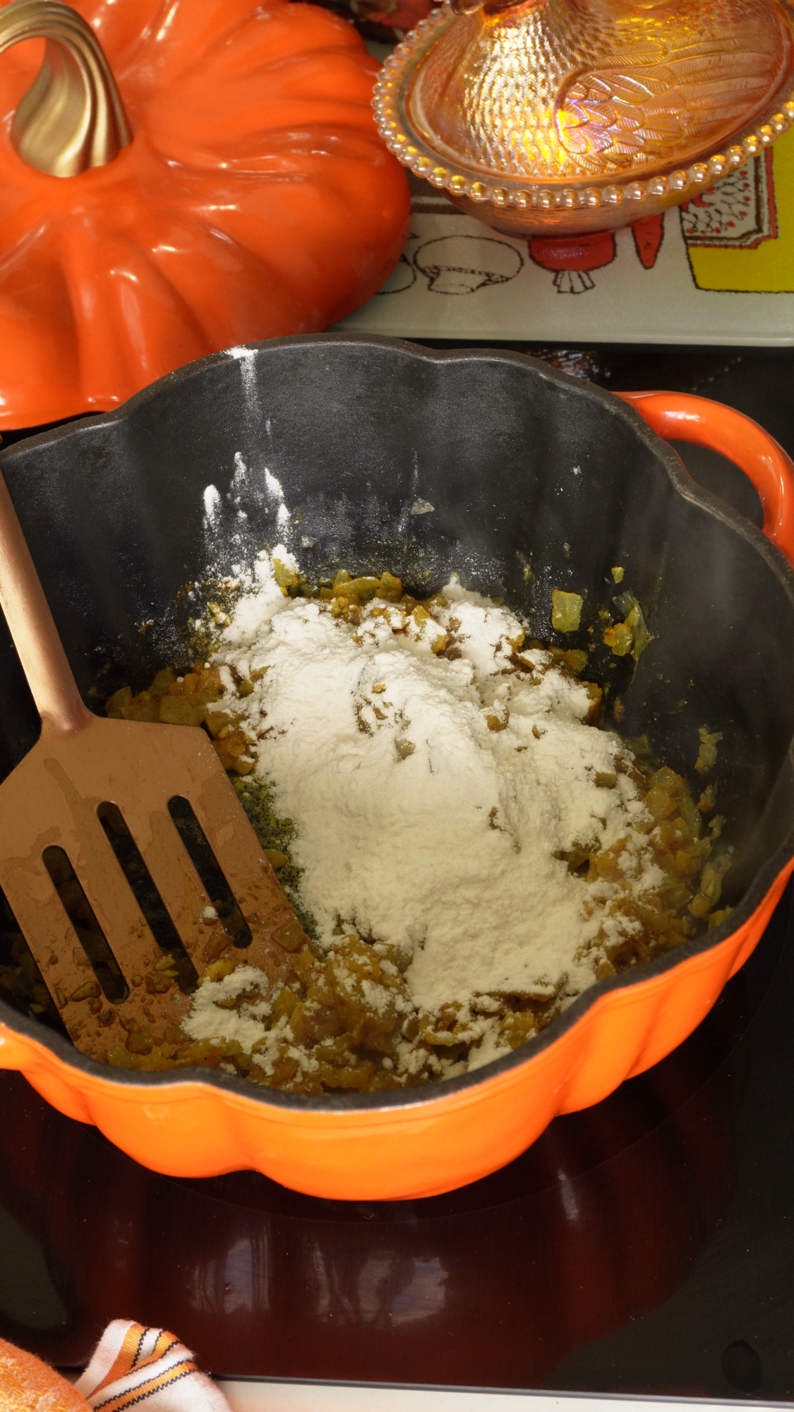 Orange pumpkin-shaped pot on a stove with chopped onions, green peppers, and flour being stirred using a spatula. A matching orange lid is nearby.