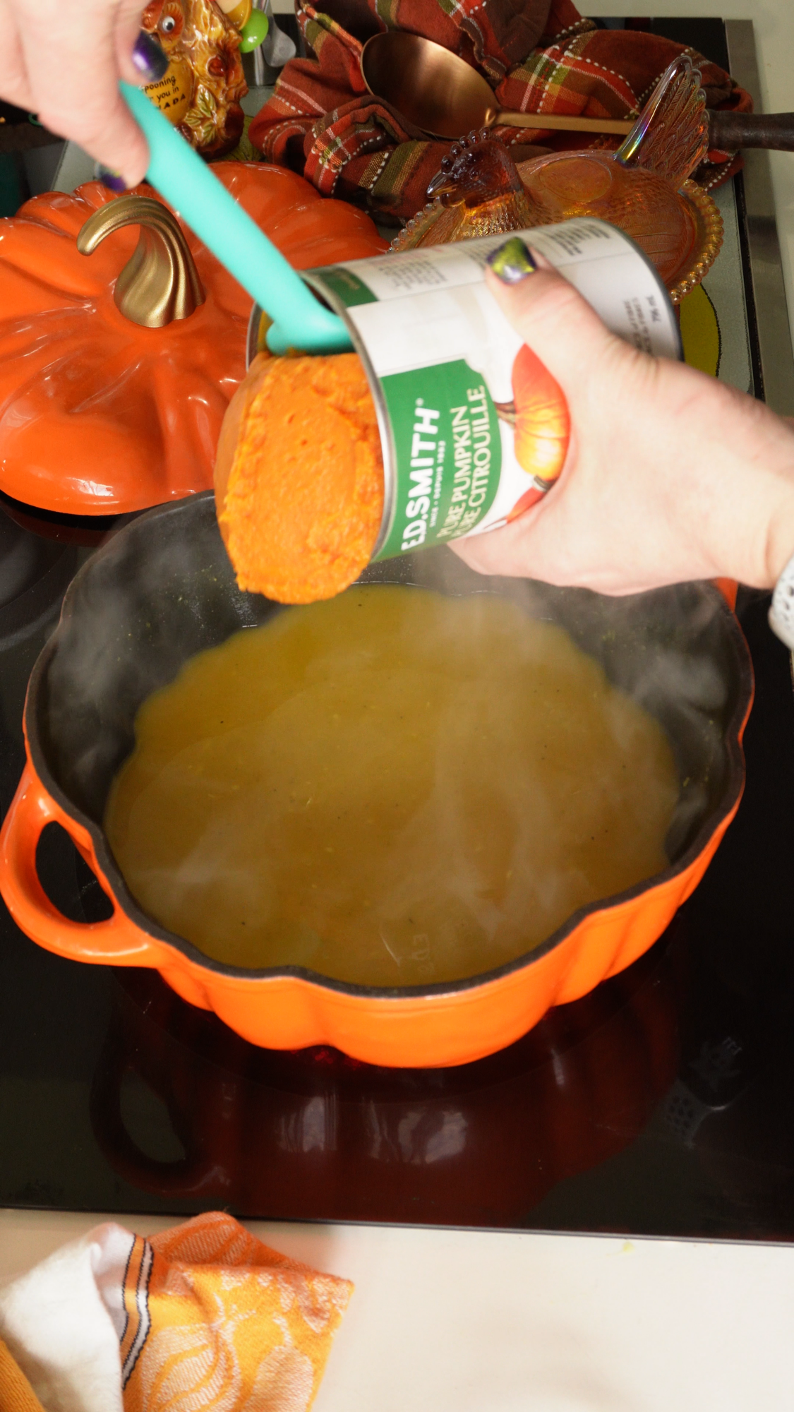 A person uses a spatula to scoop canned pumpkin puree into a steaming orange pumpkin-shaped pot on a stovetop, with autumn-themed decor in the background.