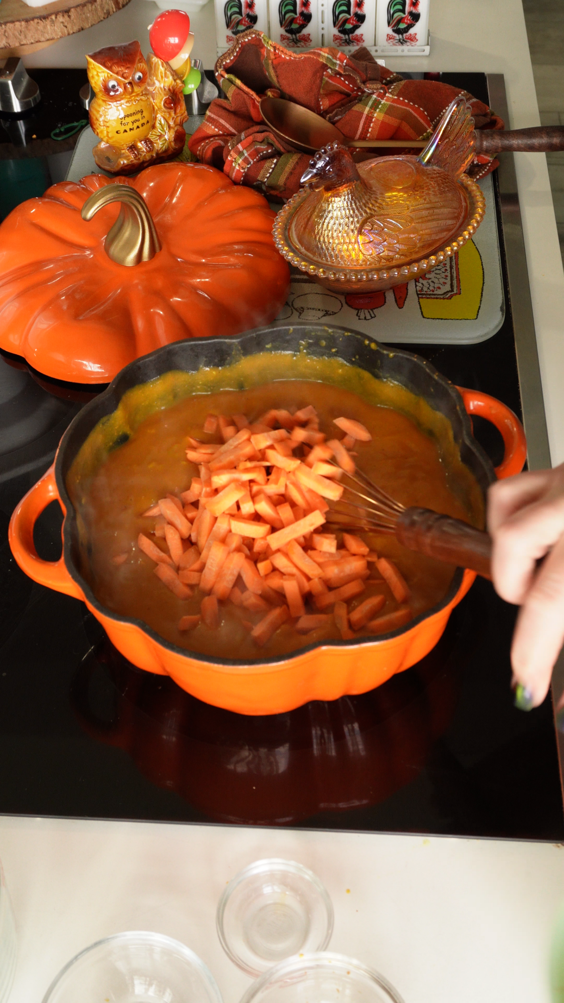 A hand stirs chopped carrots into an orange pot of soup on a stovetop, with pumpkin-themed decor and kitchen items nearby.