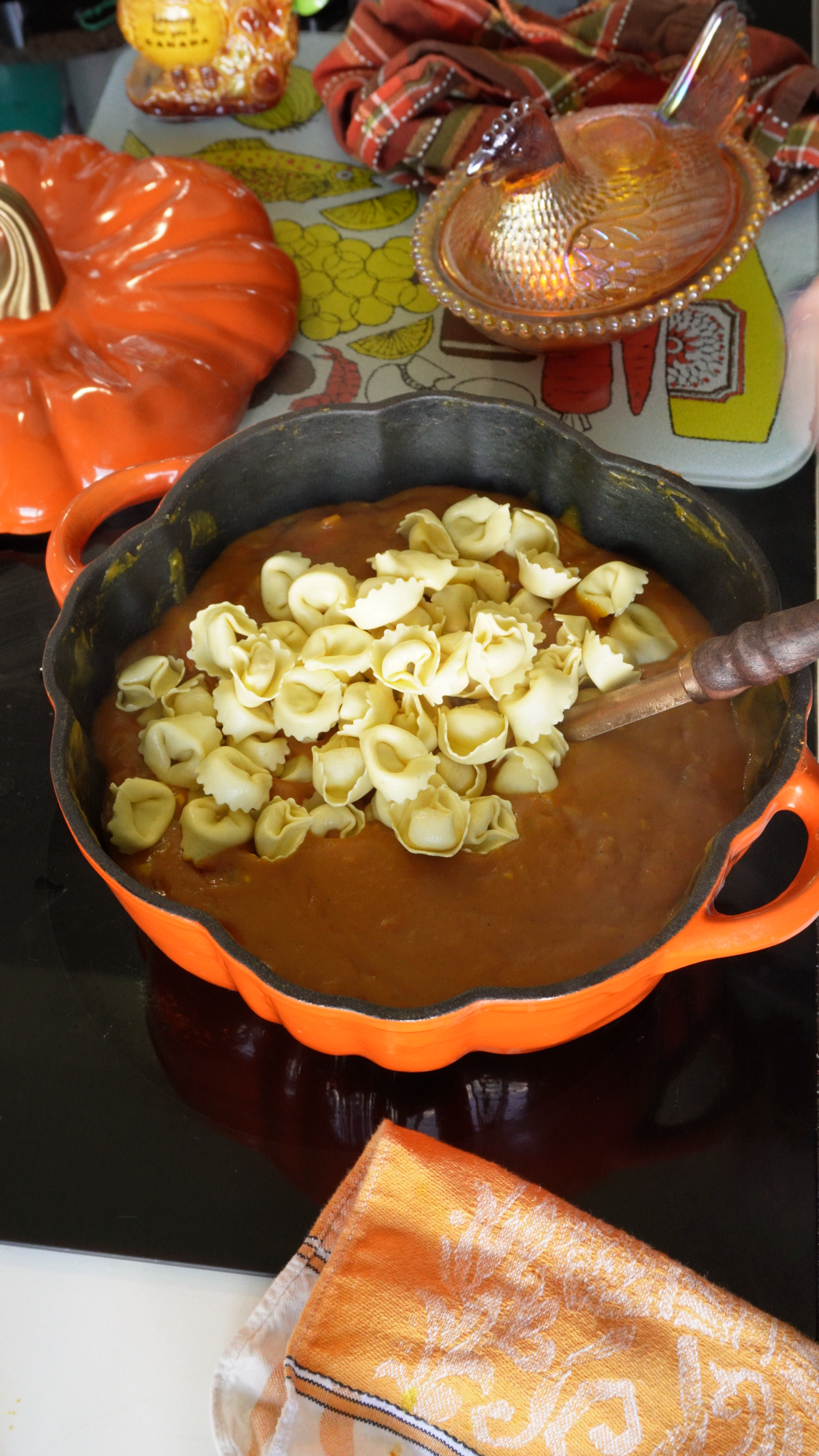 Orange pot with pasta shells being stirred into a creamy soup on a stovetop. Nearby are a pumpkin-shaped dish, napkins, and a honeycomb-patterned dish on a table with patterned textiles.