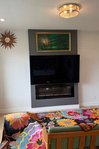 A modern living room with colorful, floral crochet blankets on furniture, a wall-mounted TV above a fireplace, a green painting, a starburst clock, and large window letting in natural light.