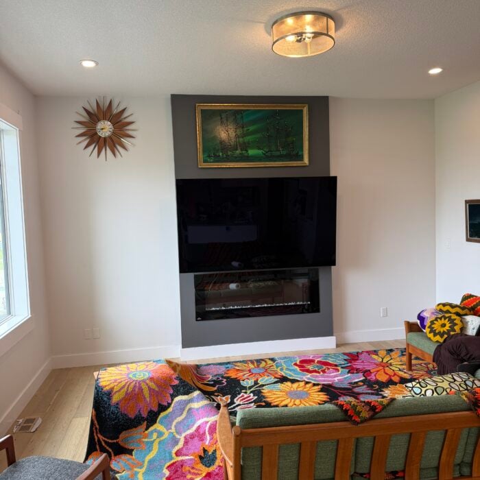 A modern living room with colorful, floral crochet blankets on furniture, a wall-mounted TV above a fireplace, a green painting, a starburst clock, and large window letting in natural light.