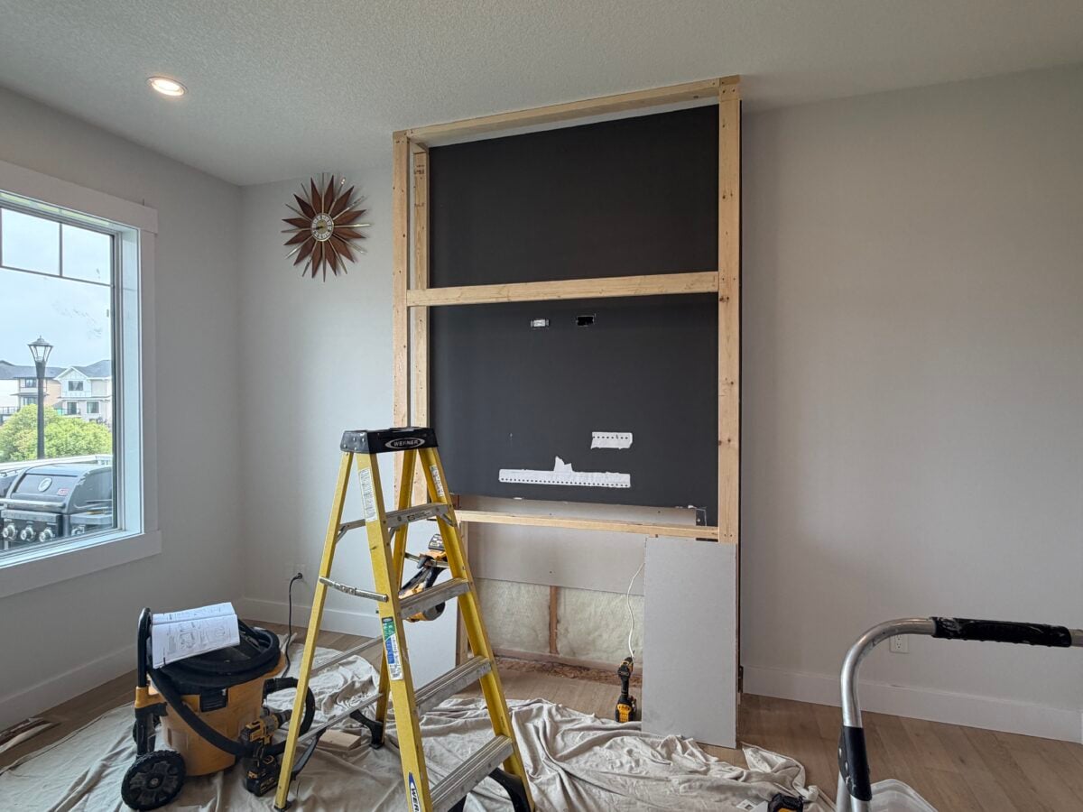 A partially built wall frame with exposed wood studs and black backing stands in a room. A yellow ladder, vacuum, and tools are nearby, with a sunburst clock on the wall and a window showing houses outside.