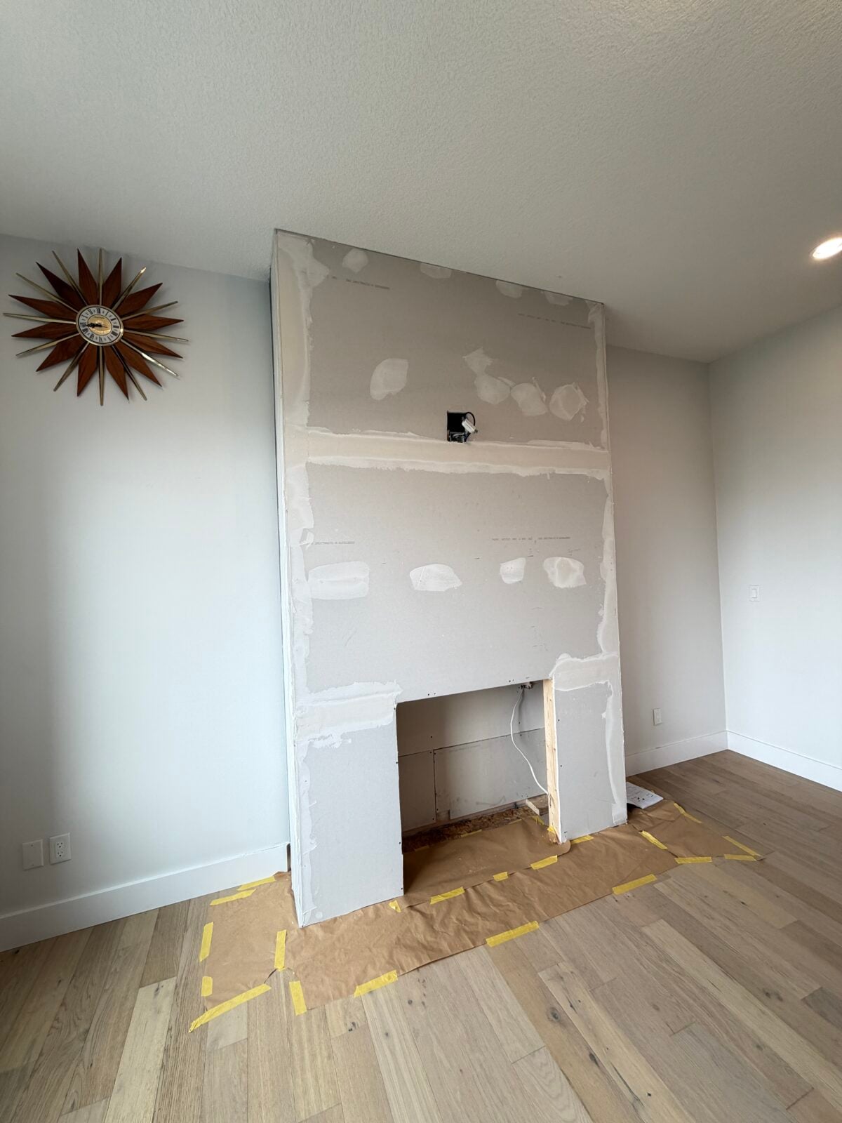 A partially constructed fireplace covered in drywall stands in a room with light wood flooring. Brown paper and yellow tape protect the floor. A mid-century modern wall clock hangs to the left.
