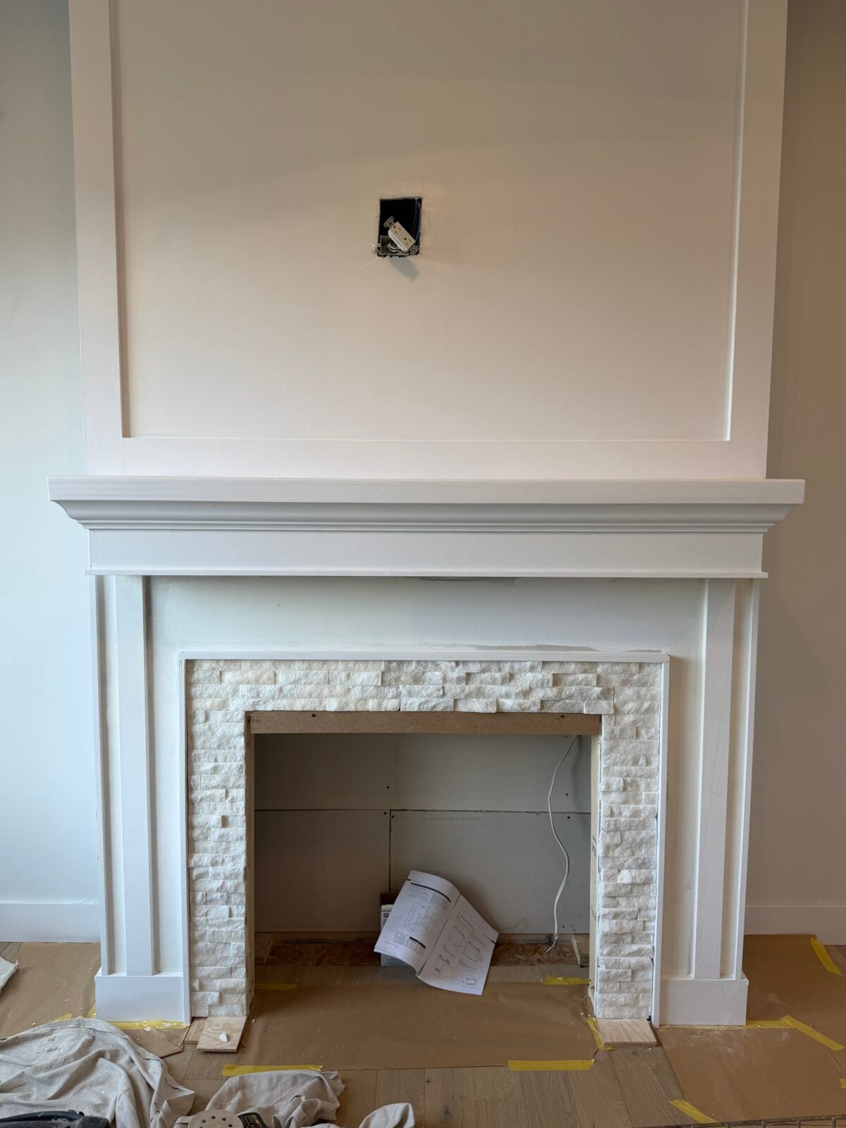 A white fireplace with a stone surround, unfinished wiring above for a possible TV or art, and some papers and cables on the bare floor in front. The area appears to be under renovation.