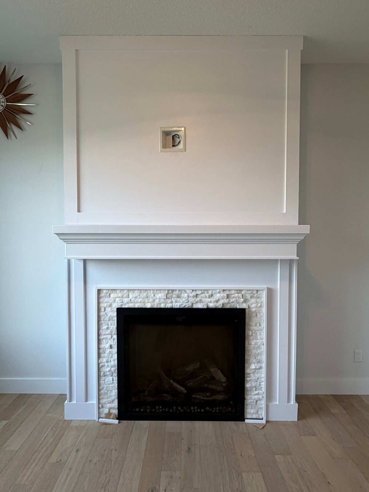 A white fireplace with a black firebox and textured stone surround stands against a light wall. Above the mantel is a small framed object. Light wood flooring is visible.