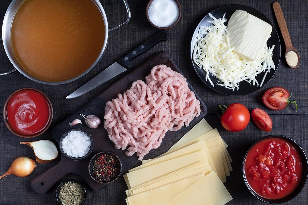 Ingredients for lasagna are arranged on a dark surface, including ground meat, lasagna noodles, tomatoes, tomato sauce, cheese, broth, spices, an onion, garlic, and a knife.