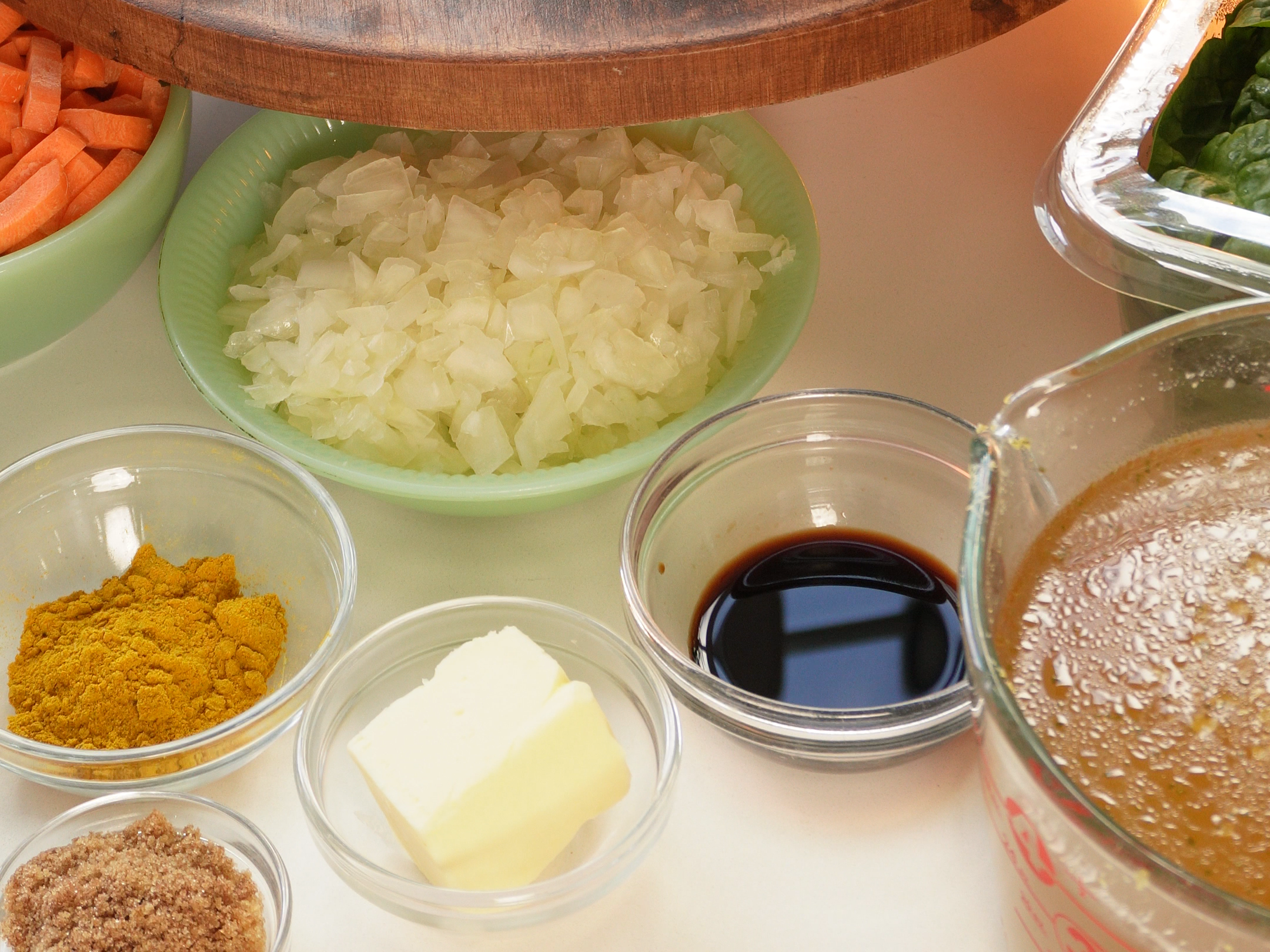 A wooden tray holds a leaf-patterned box, a green glass hen, an orange candle holder, tortellini, chopped onions, carrots, spices, soy sauce, butter, and fresh greens, with flowers in the background.