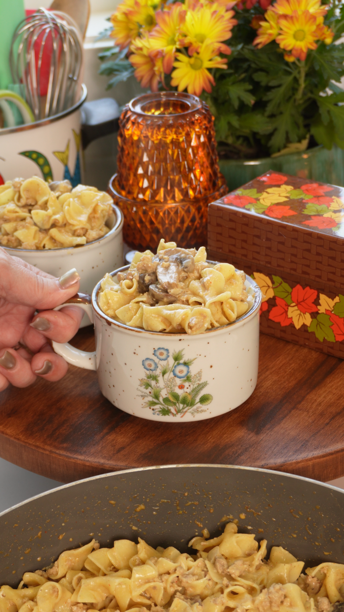 A hand holds a ceramic mug filled with beef stroganoff, with a second mug and a skillet of the same pasta visible nearby.