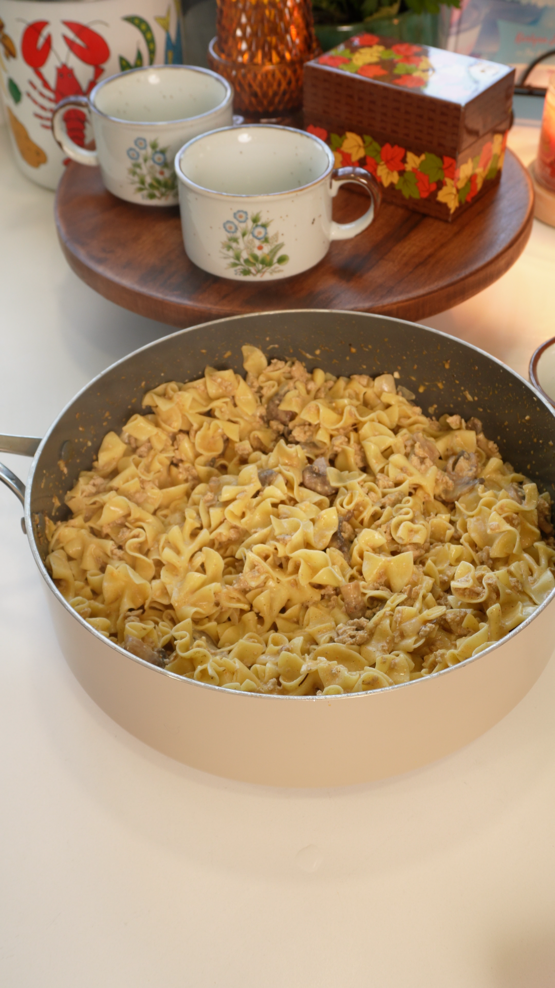 A large pan filled with creamy egg noodles and ground meat, with two floral mugs and a patterned box on a wooden board in the background.
