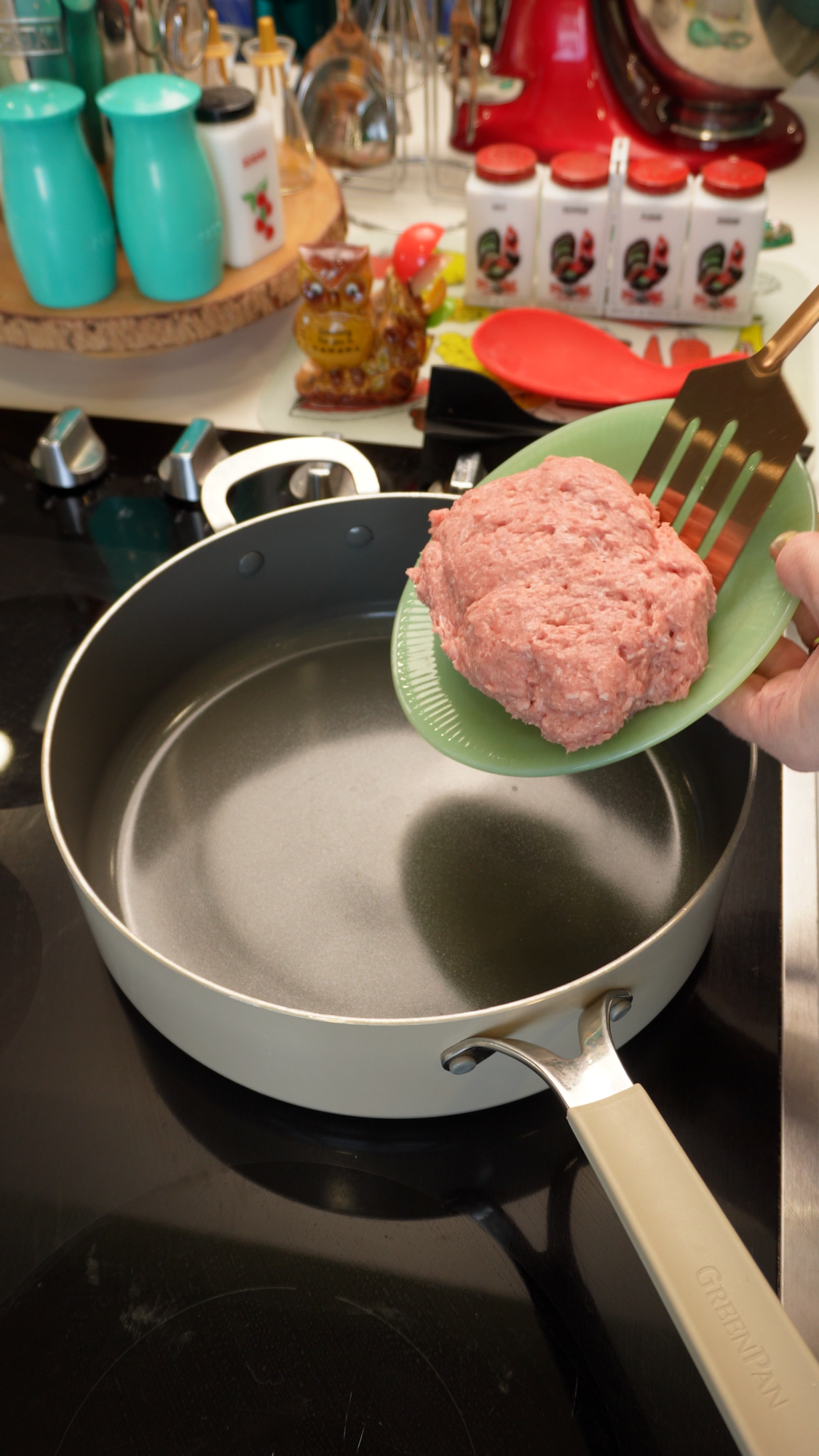 A hand uses a green slotted spatula to place a mound of ground meat into a light-colored nonstick skillet on a stovetop.