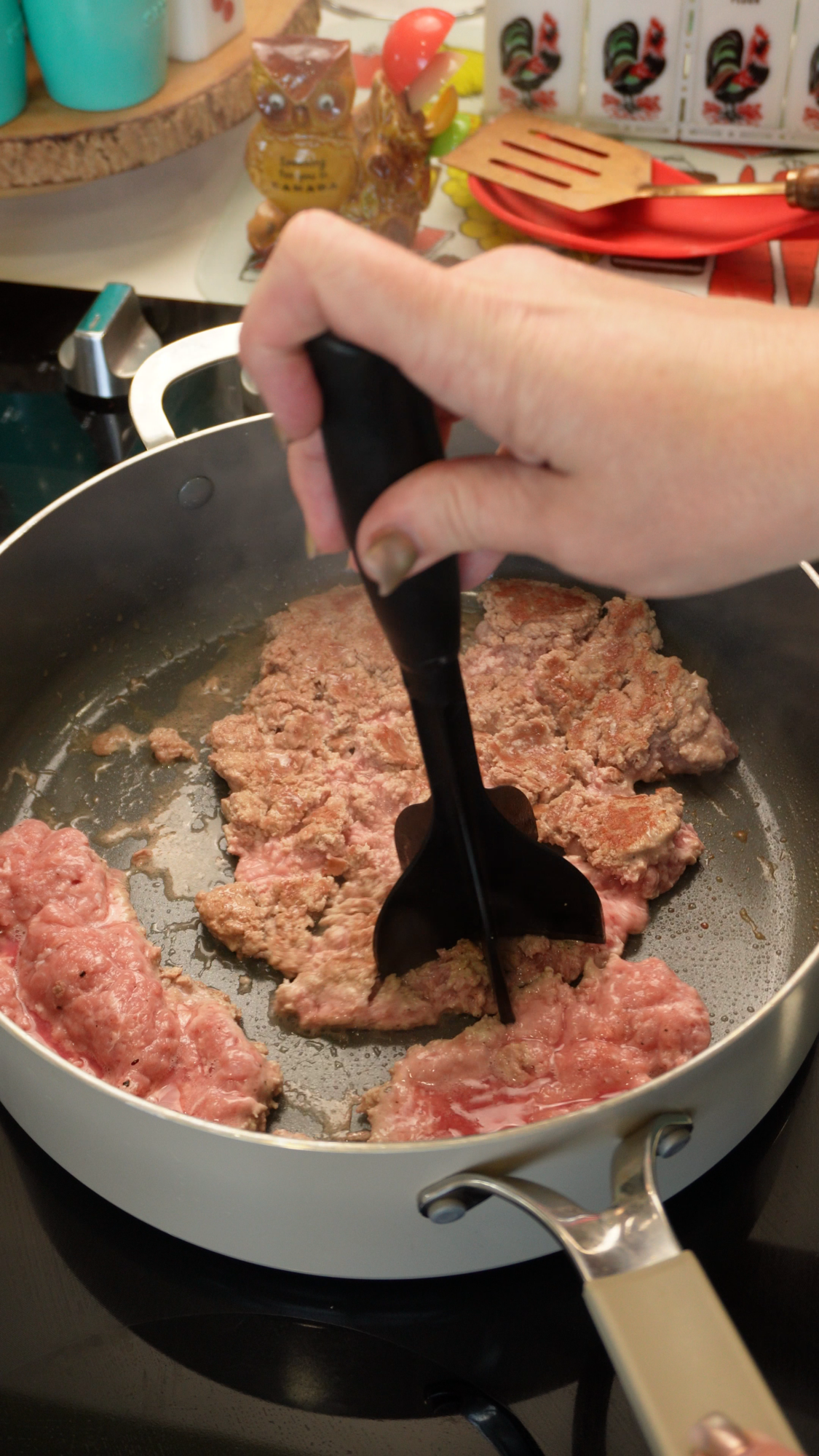 A hand uses a black meat masher to break up ground meat browning in a pan.