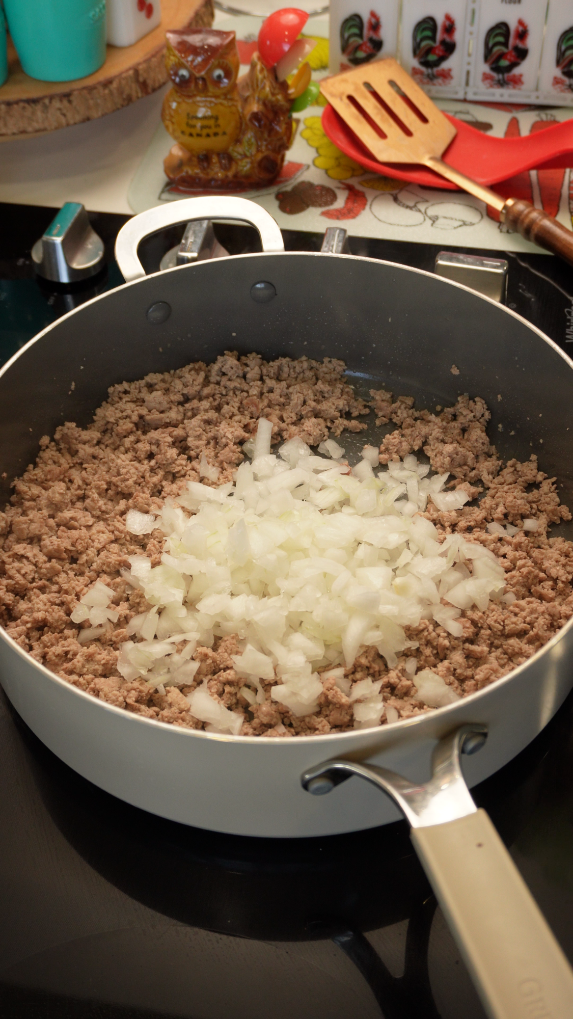 A pile of diced white onions rests on top of browned ground meat in a skillet on a stovetop.