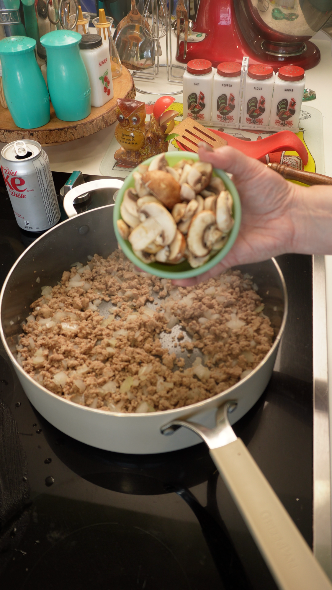 A hand holds a small green bowl of sliced mushrooms over a pan containing ground meat and diced onions.