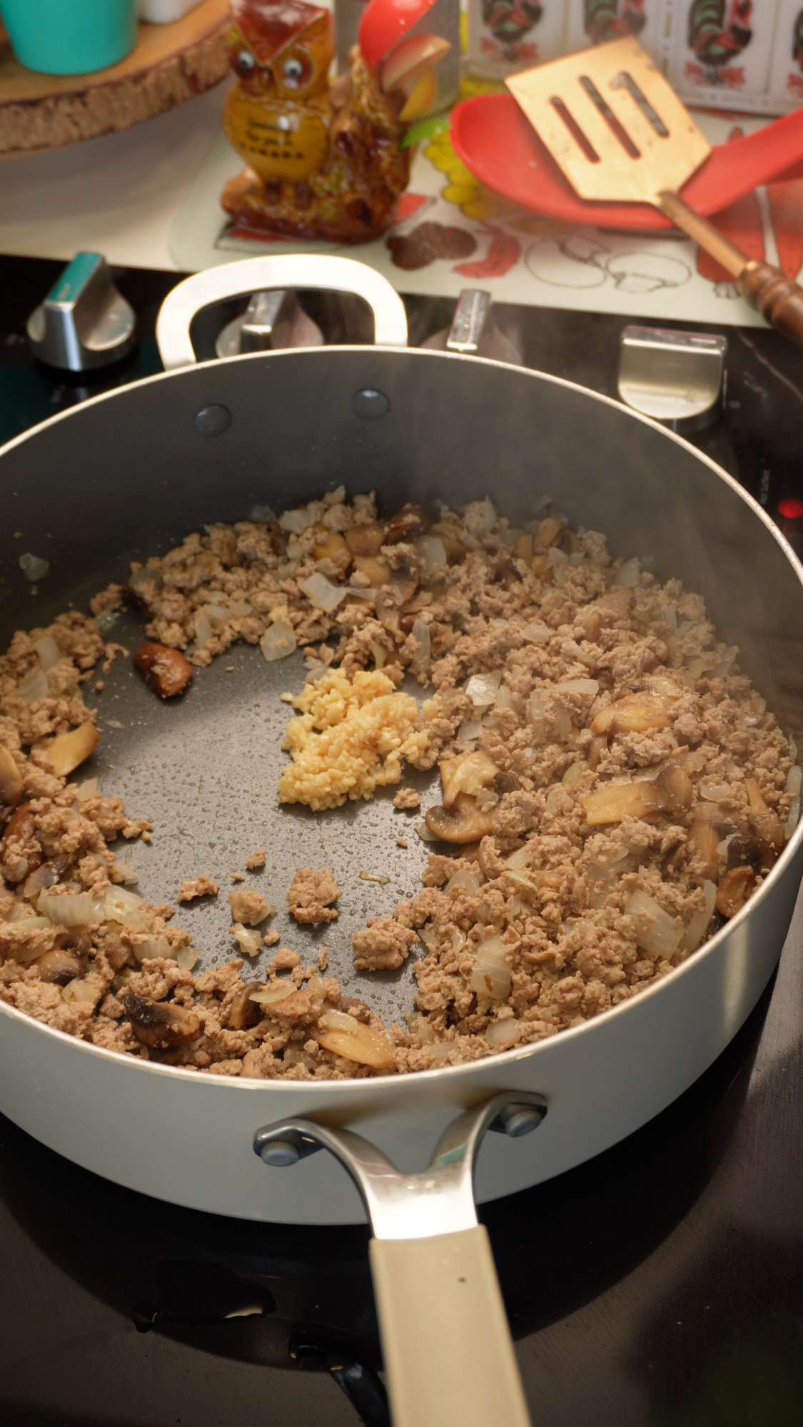 Ground meat, chopped onions, and sliced mushrooms cooking in a pan with a scoop of minced garlic in the center.