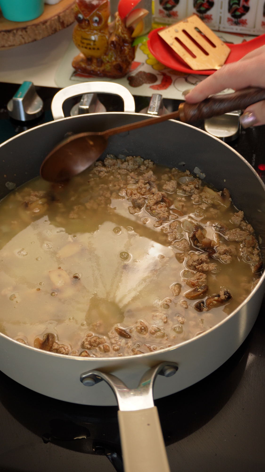 A person uses a wooden spoon to stir a mixture of ground meat and sliced mushrooms simmering in a light-colored pan on a stovetop.
