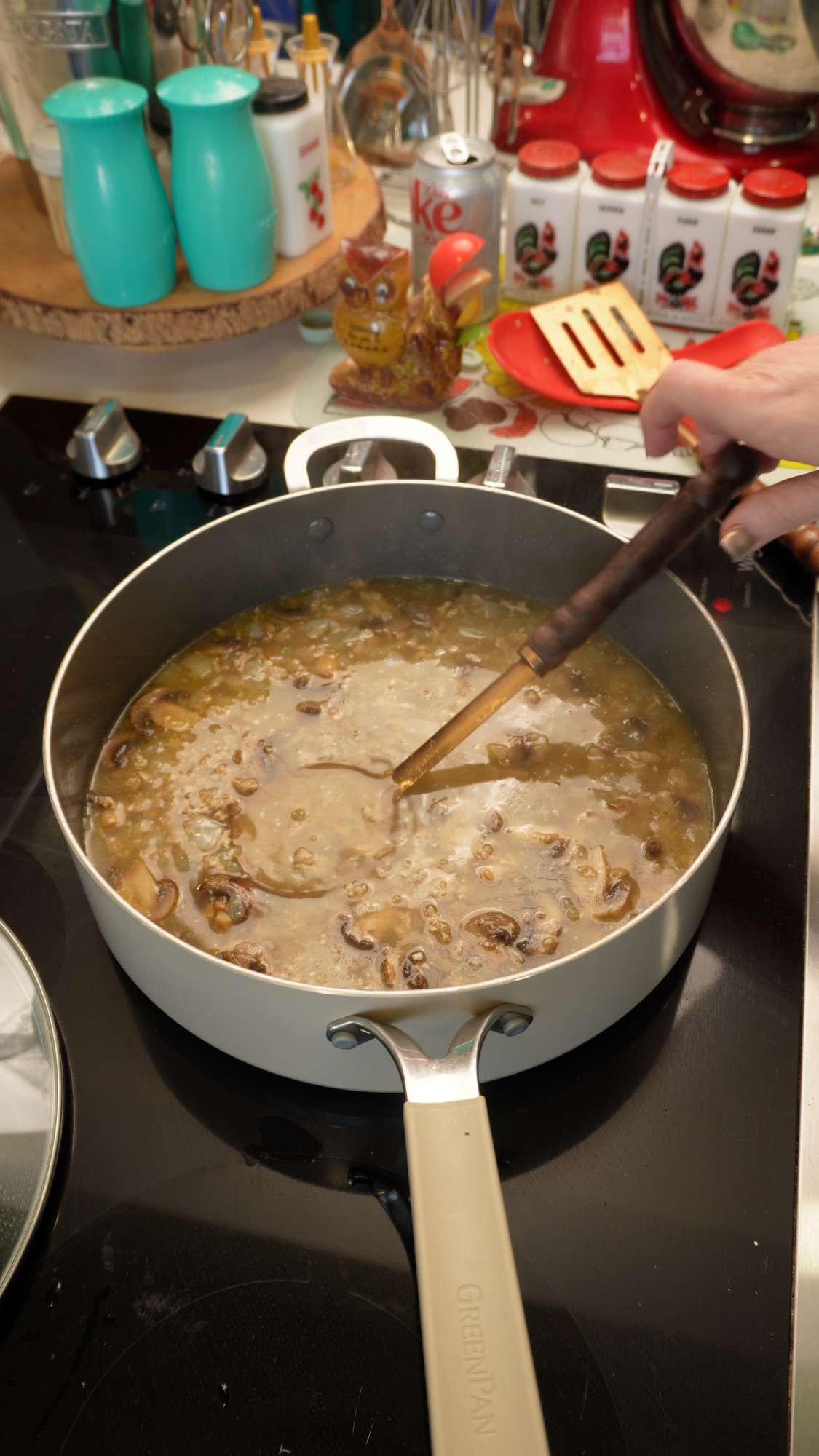 A hand uses a ladle to stir a mushroom soup in a large, light-colored pan on a black stovetop.