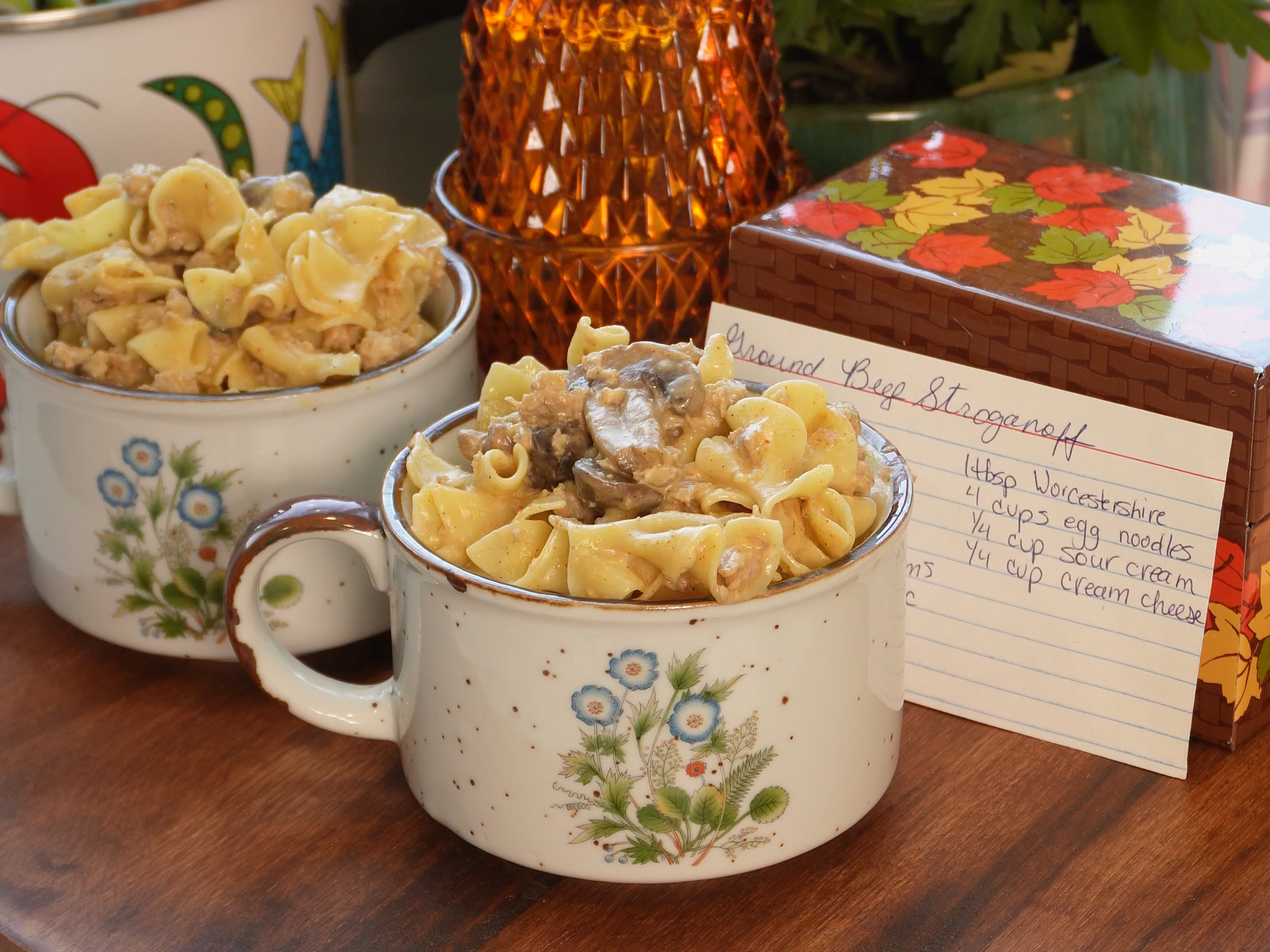 Two mugs filled with beef stroganoff sit on a wooden table next to a handwritten recipe card.