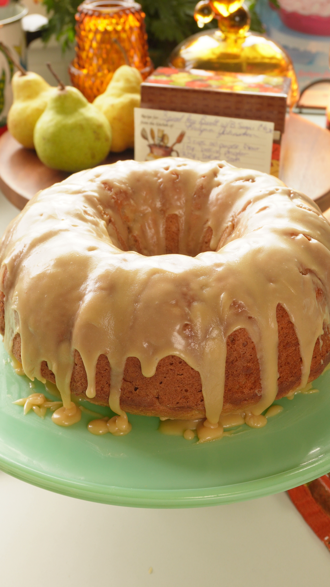 A bundt cake with caramel-colored glaze on a green pedestal stand, with two pears in the background.