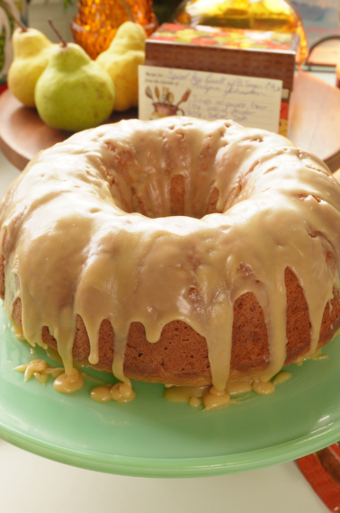 A bundt cake with caramel-colored glaze on a green pedestal stand, with two pears in the background.