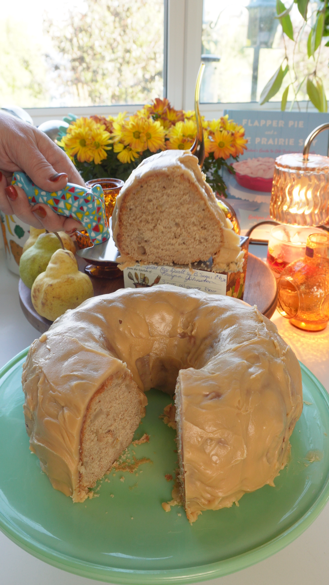 A sliced bundt cake with caramel frosting on a green cake stand, with a slice held by a hand and pears in the background.