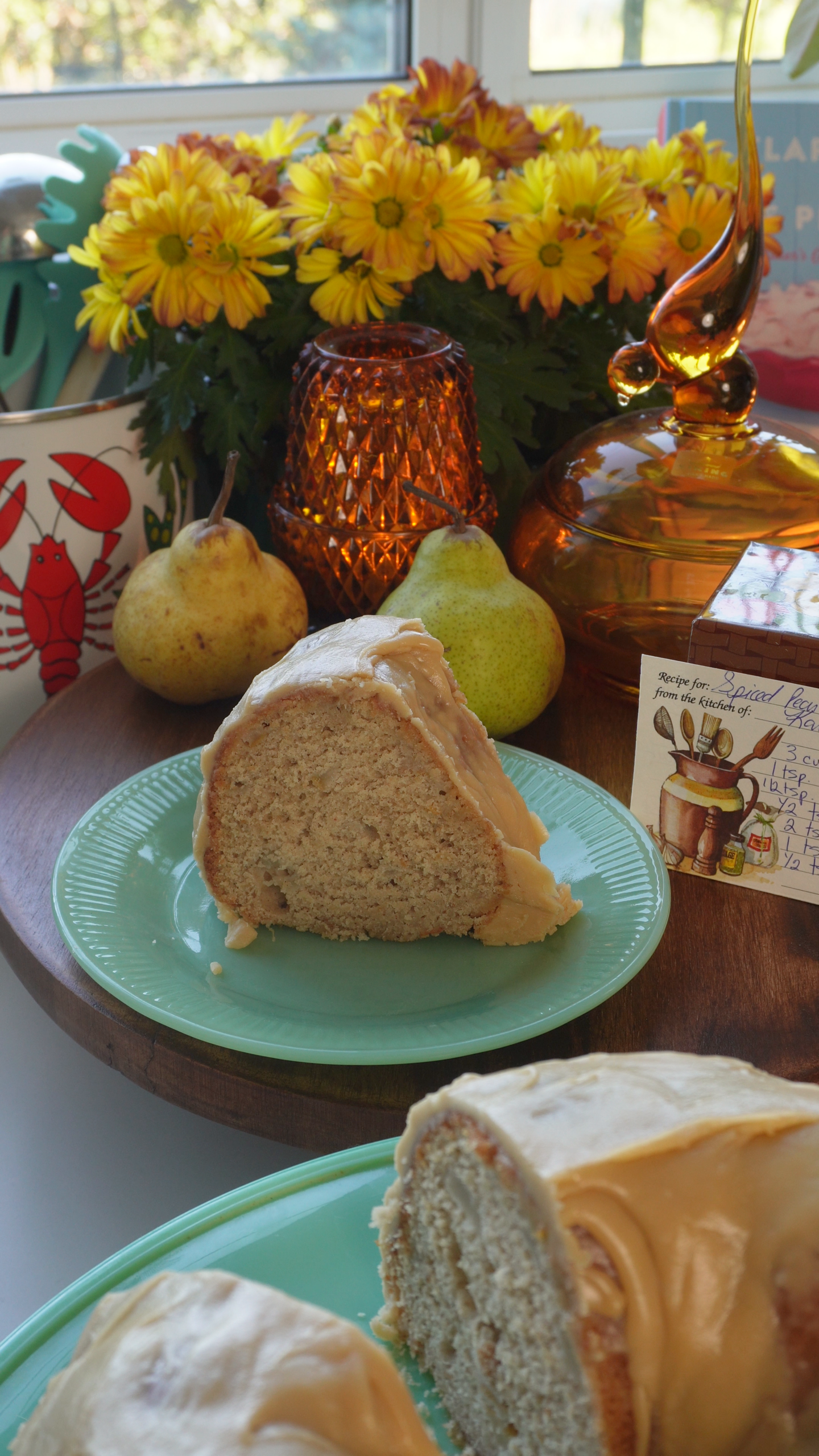A slice of glazed spice cake on a light green plate, with two pears, yellow flowers, and a handwritten recipe card in the background.
