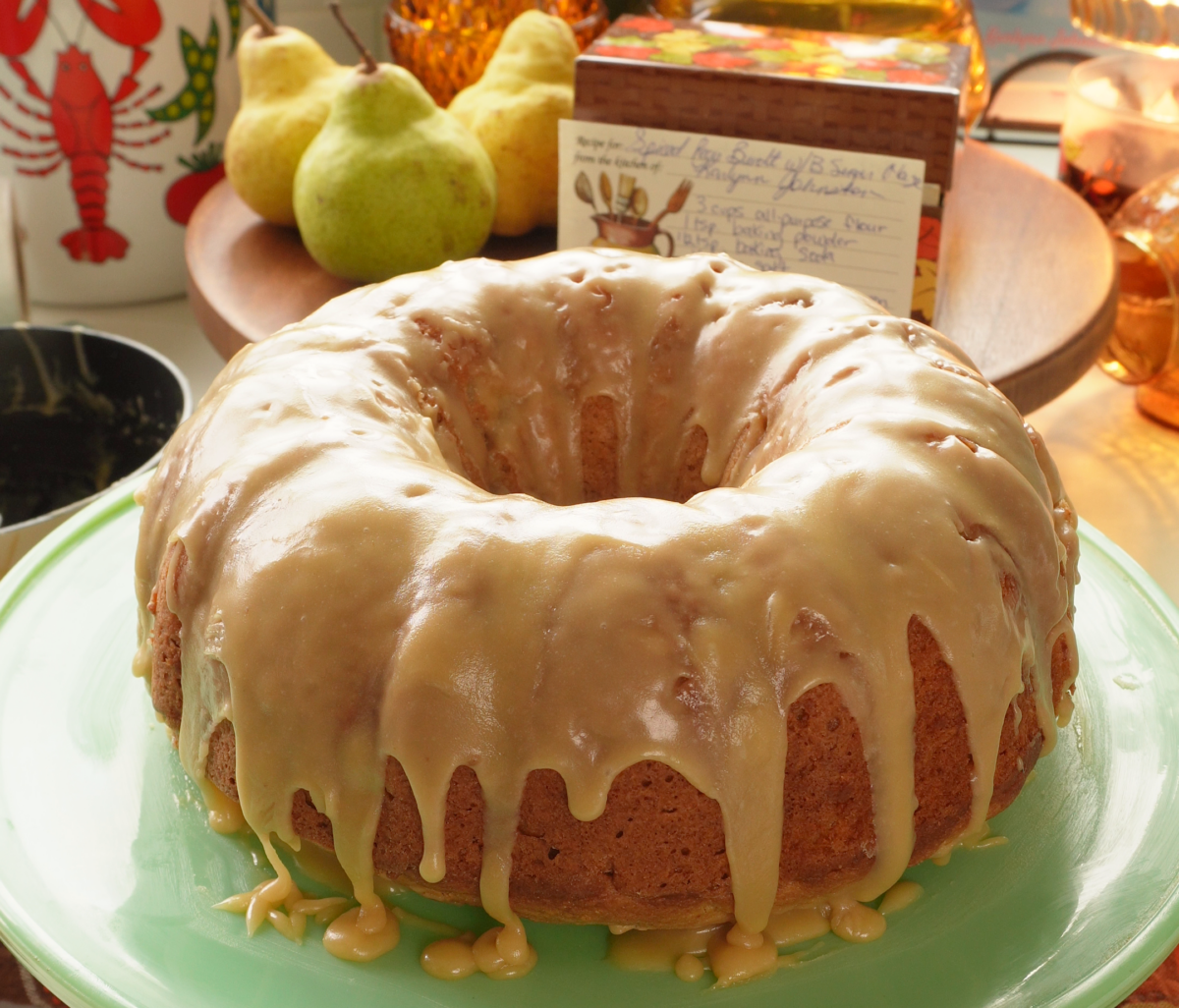 A glazed bundt cake sits on a light green plate, with pears and a handwritten recipe card in the background.