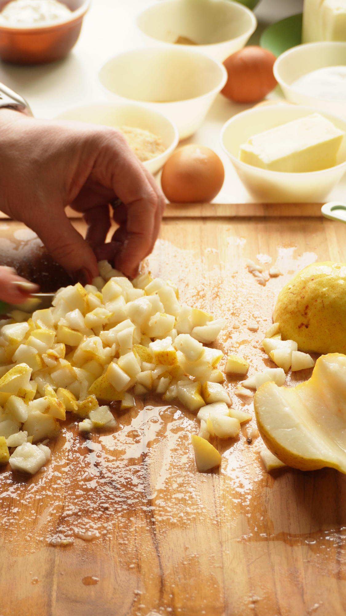 Hands chopping a pear into small cubes on a wooden cutting board, with bowls of baking ingredients in the background.