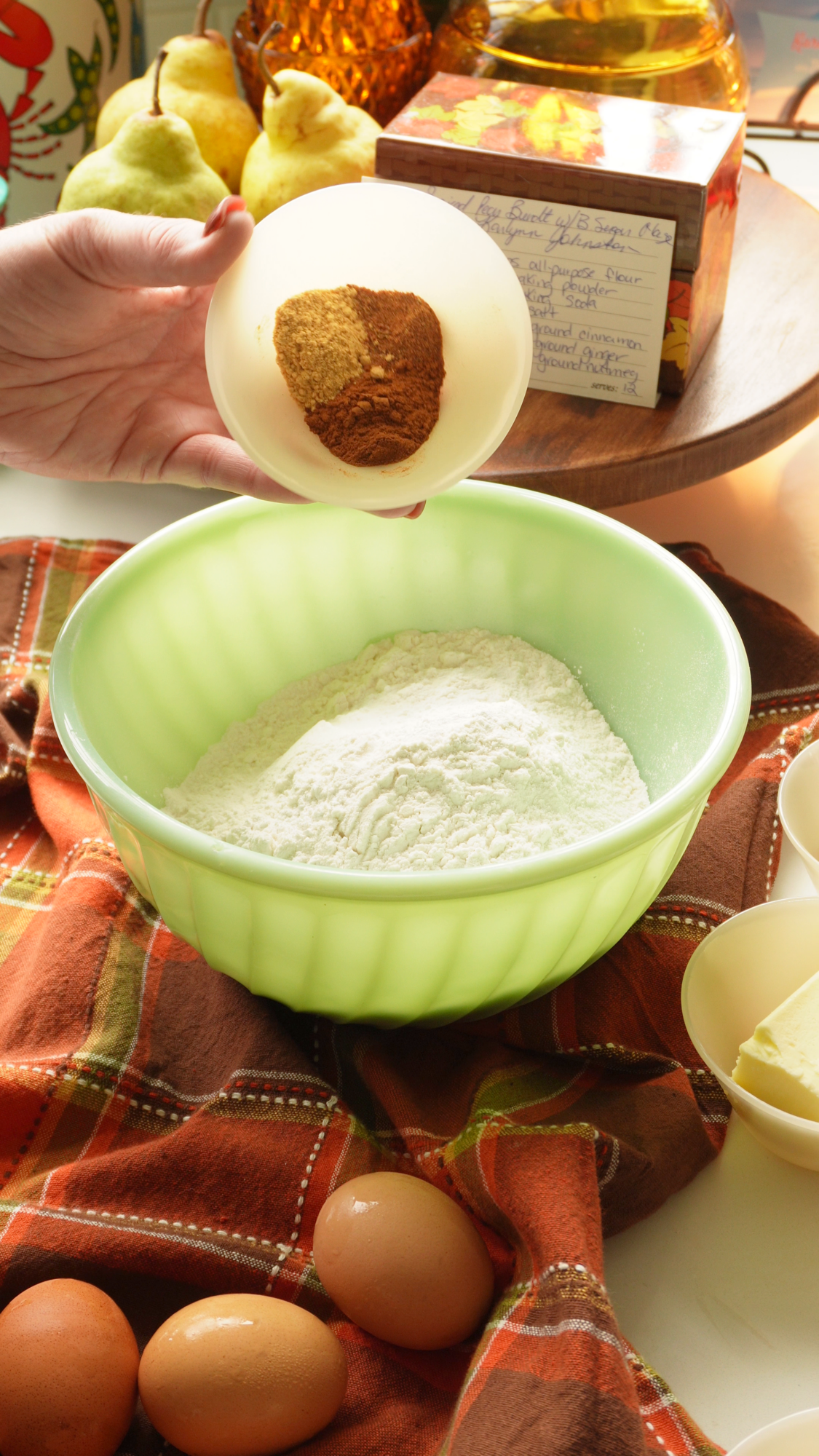 A hand holds a small bowl containing ground spices over a large mint-green bowl filled with flour.