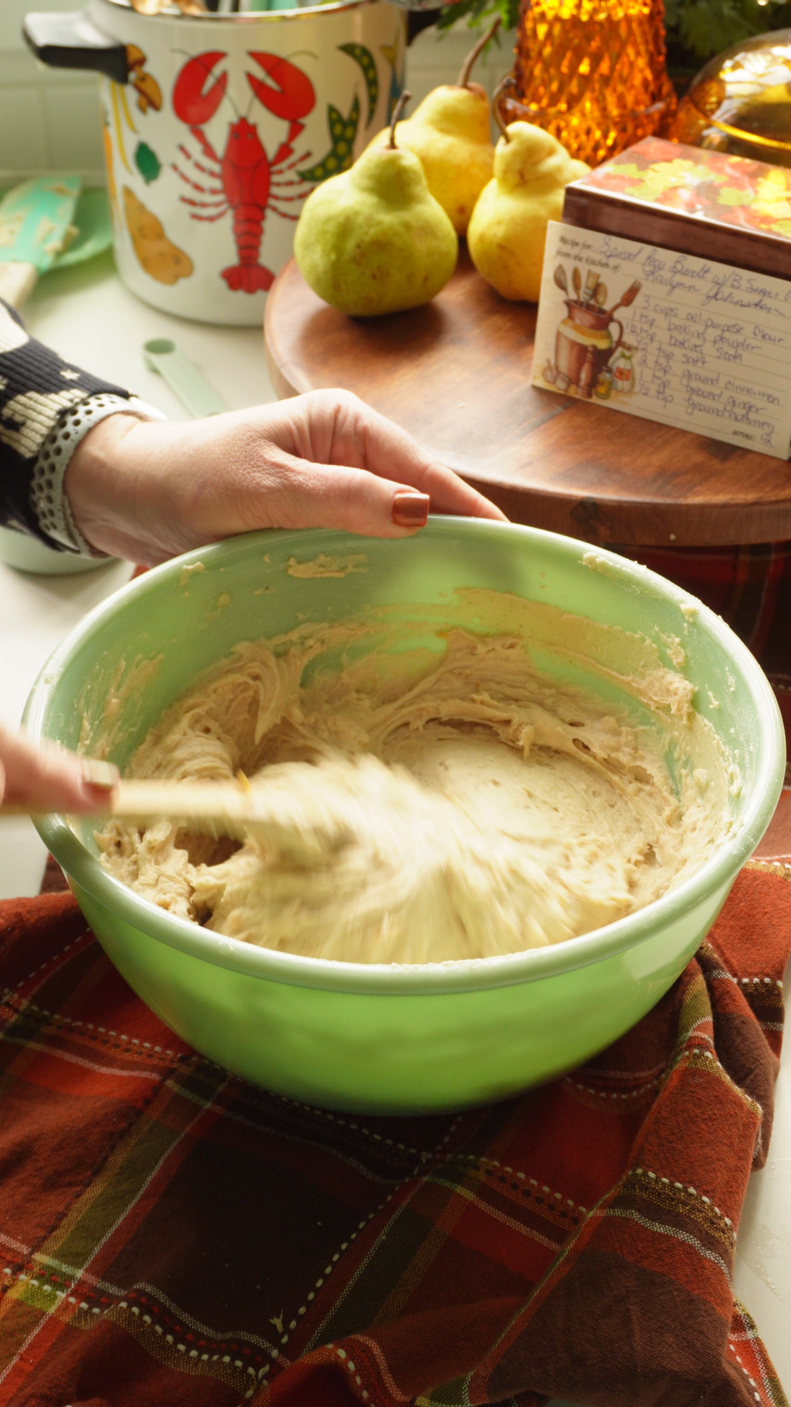 A hand uses a spatula to mix batter in a light green bowl, with pears and a recipe card on a wooden board in the background.
