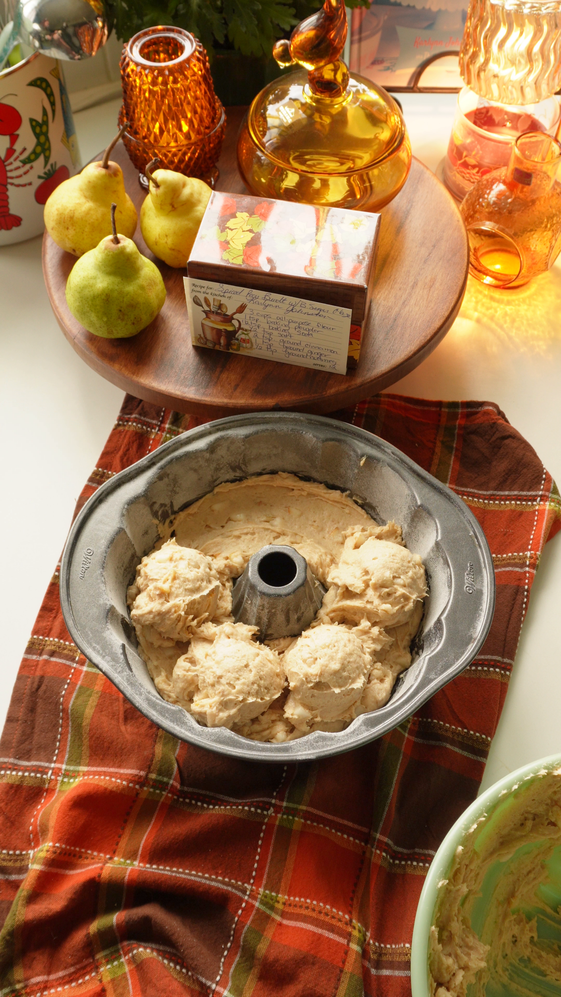 Bundt pan filled with scoops of cake batter sitting on a plaid cloth, with a recipe card and pears on a wooden riser in the background.