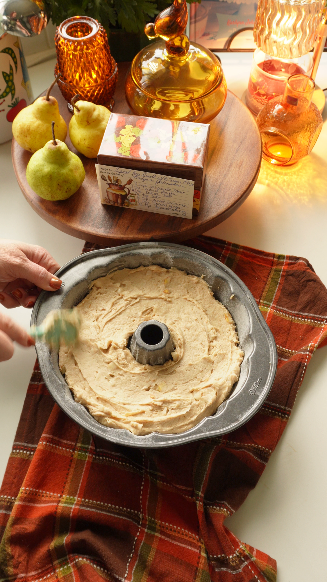 A hand uses a spatula to spread thick cake batter into a bundt pan, with fresh pears and a handwritten recipe card on a wooden stand in the background.