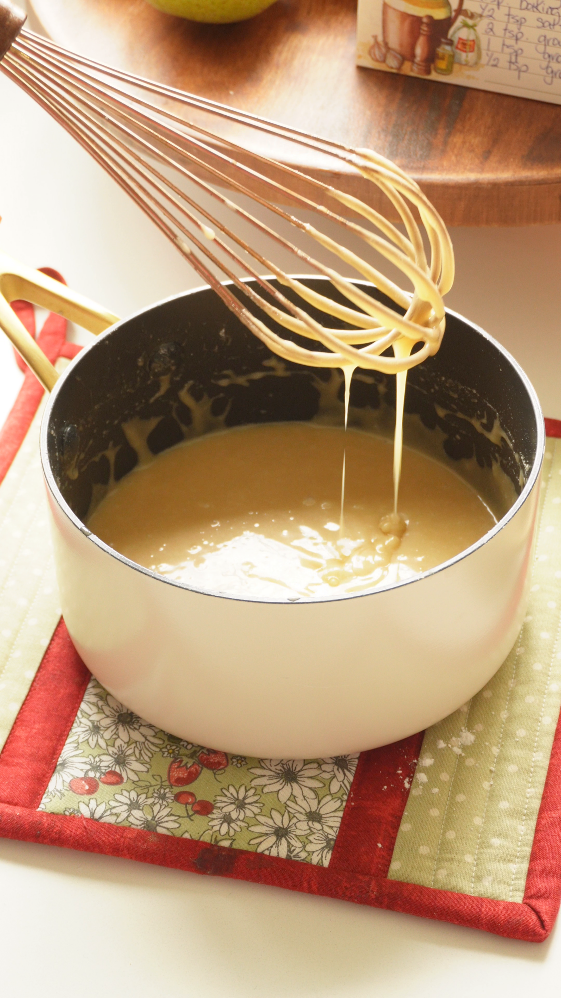 A whisk drips batter into a white saucepan sitting on a quilted pot holder.