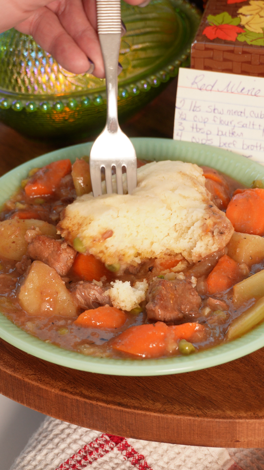 A fork lifts a piece of crust from a green bowl of beef stew containing carrots, potatoes, and peas, with a handwritten recipe card in the background.