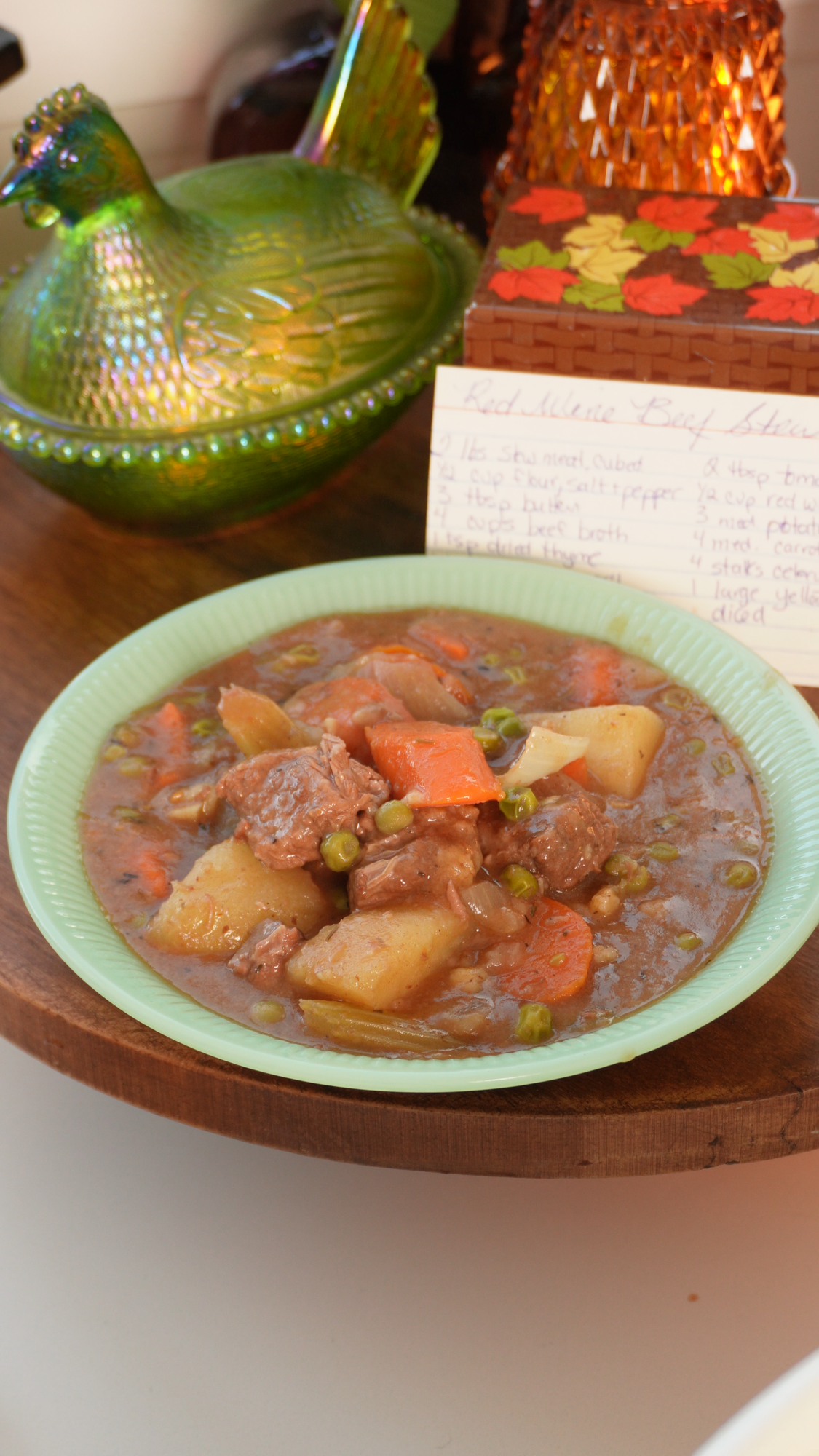 A bowl of beef stew with potatoes, carrots, and peas on a wooden tray next to a handwritten recipe card and a glass hen dish.