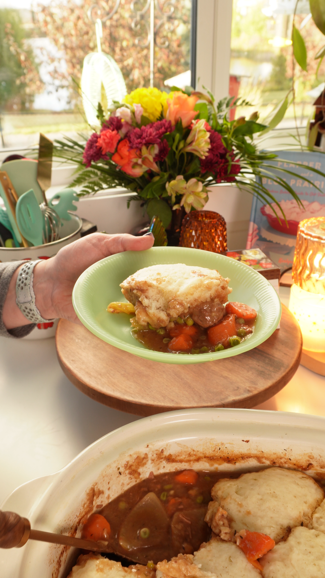 A hand holds a plate containing a serving of beef stew topped with a biscuit, with a larger casserole dish of the same meal in the foreground.