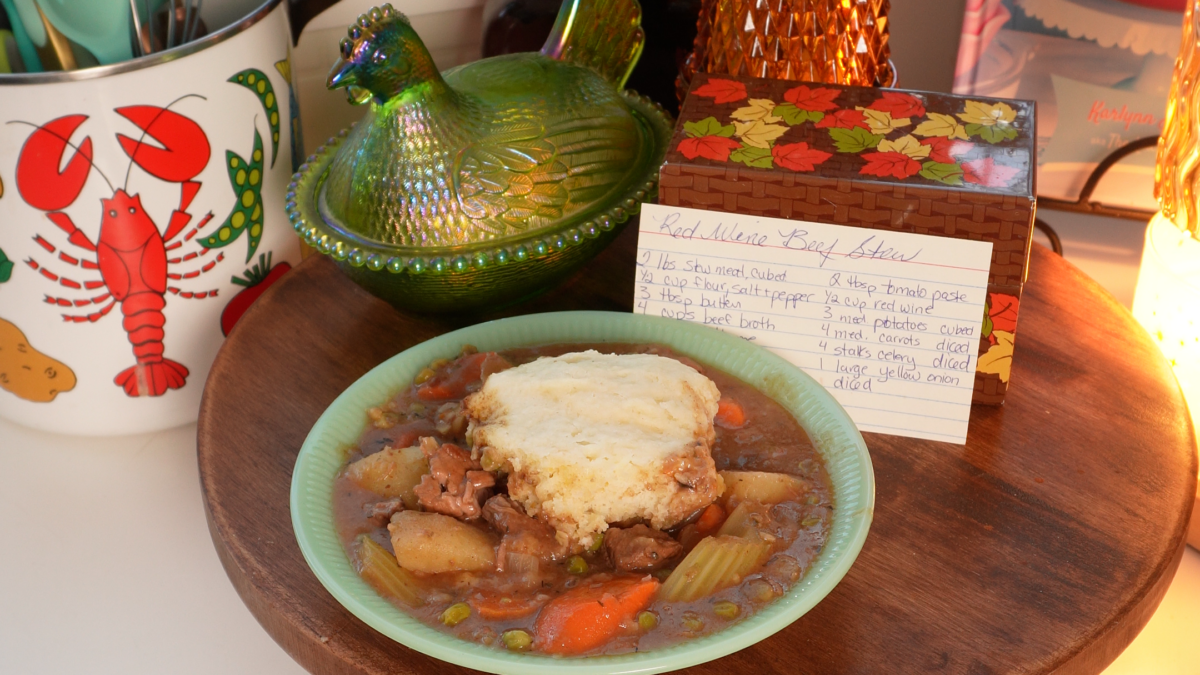 A bowl of beef stew topped with a biscuit, sitting on a wooden surface next to a handwritten recipe card for Red Wine Beef Stew.